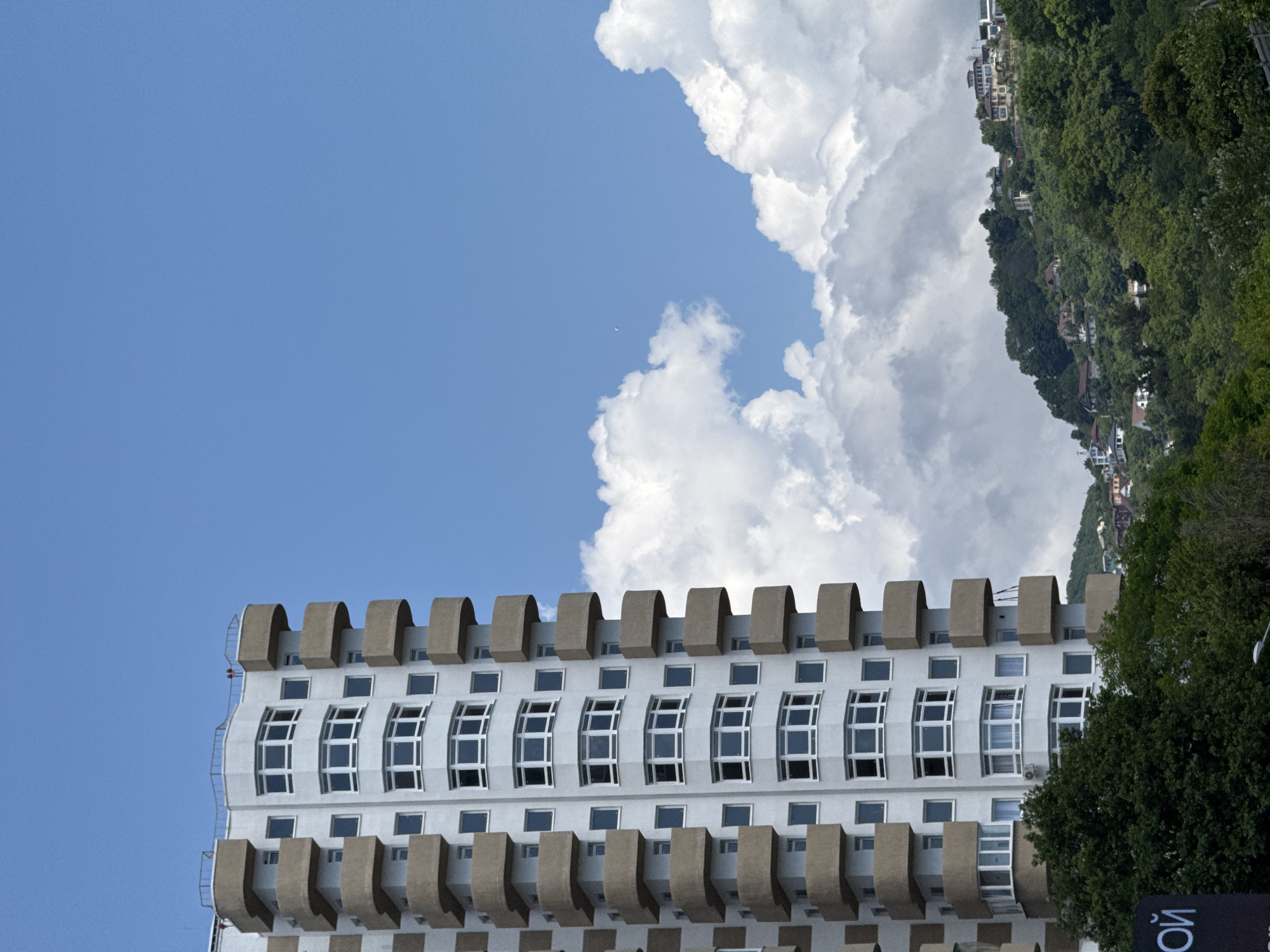 The image depicts a modern residential building with numerous balconies, set against a backdrop of a lush green hillside dotted with houses. The sky is bright blue with fluffy white clouds. The building is tall and white with beige trim on the balconies. The hillside appears to be a residential area with buildings of varying architectural styles. The overall scene suggests a sunny day in a city with a mountainous landscape. The text on the black sign at the bottom left of the image appears to be in Russian. While the specific location in Sochi, Russia, is not identifiable from the image alone, the presence of the Russian text and the general aesthetic suggest it could be a coastal city on the Black Sea.