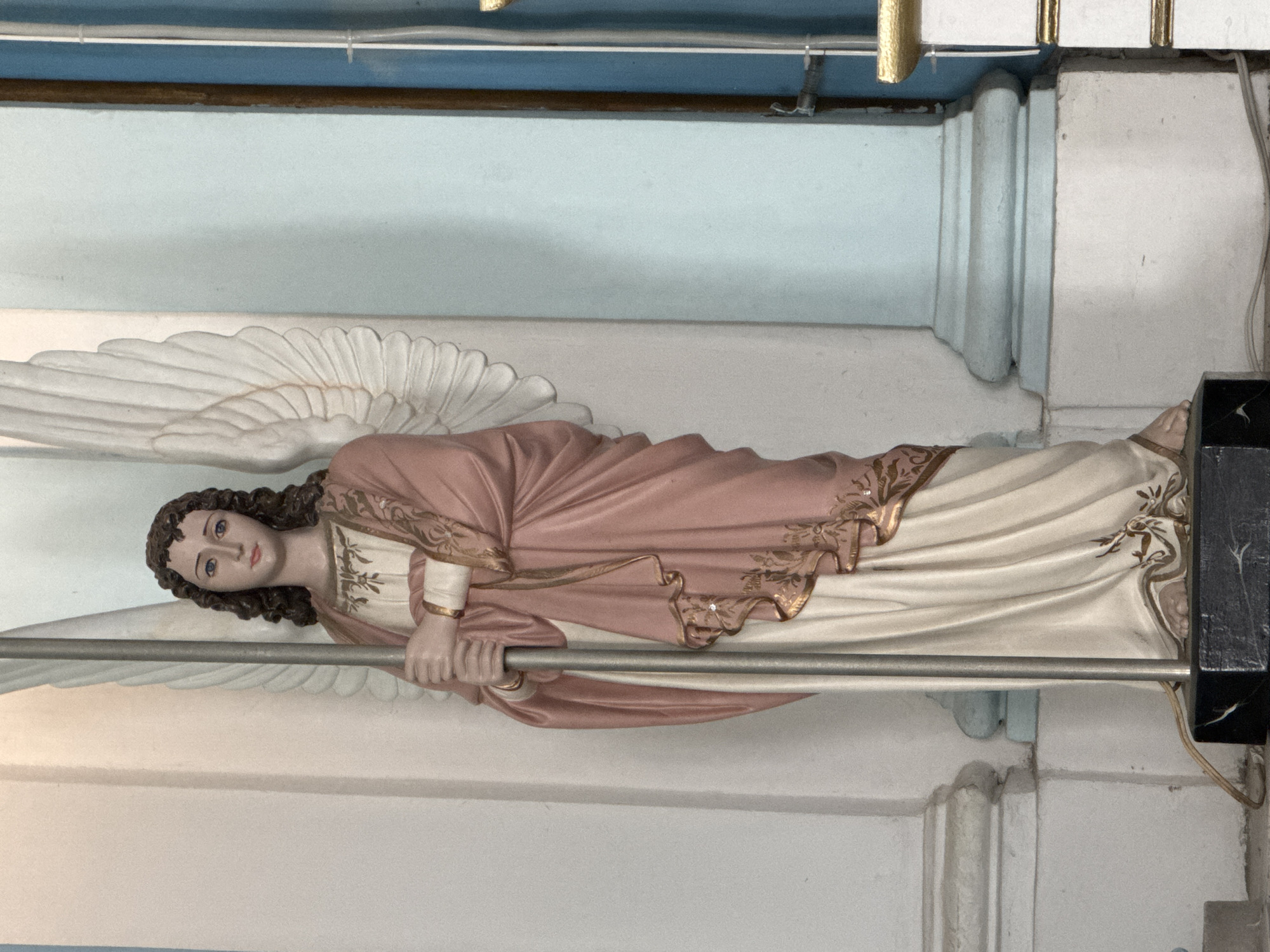 The image shows a full-body statue of an angel, likely made of plaster or resin, standing in what appears to be a church or religious building. The statue is positioned in front of a white pillar on the left and a light blue wall on the right. The angel has dark curly hair, blue eyes, and is depicted with a serene expression. It is wearing a long, flowing cream-colored robe adorned with gold trim at the hem, and a draped pink mantle with intricate gold embroidery. A gold band encircles each wrist. The angel holds a long, slender silver staff vertically in front of its body with both hands. Large white wings extend from its back, with the right wing prominently visible and the left wing partially obscured by the staff and body. The statue stands on a black rectangular base. The background consists of architectural elements typical of a sacred space, including additional pillars and walls. The lighting suggests an interior setting with diffused light.
This statue is located in the Igreja de São Francisco Xavier, in the district of Icaraí, Niterói, Brazil.