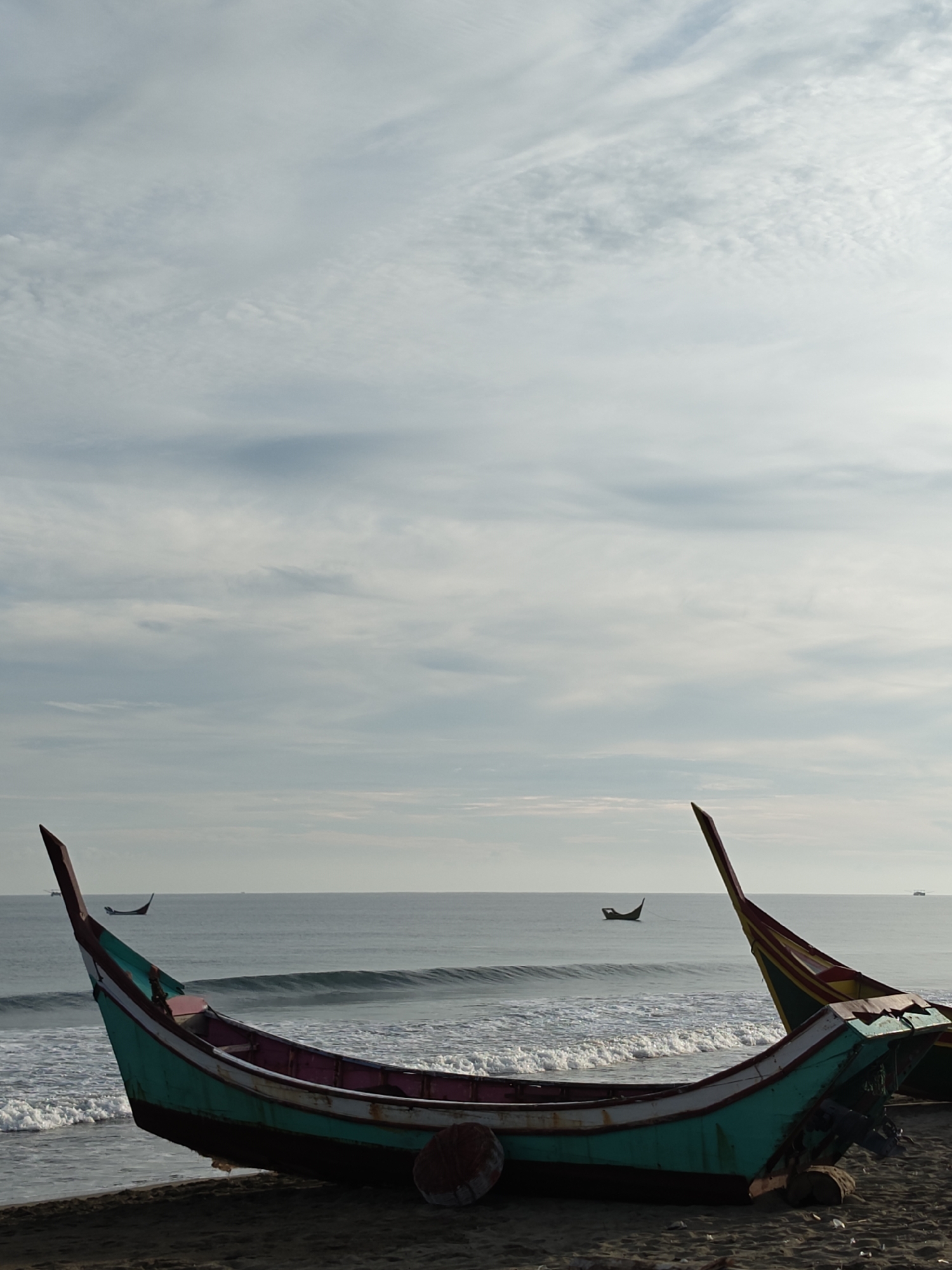 An overcast sky with wispy clouds stretches above a serene ocean and a dark sandy beach in Curehtunong, Indonesia. The foreground is dominated by the hull of a traditional Indonesian fishing boat, its vibrant teal and pink colors contrasting with the muted tones of the surroundings. The boat rests on the sand, partially submerged in the incoming tide, with gentle waves lapping at its sides. In the distance, two other smaller fishing boats are visible on the calm water, appearing as dark silhouettes against the hazy horizon. The overall atmosphere is one of quiet solitude and the gentle rhythm of coastal life.