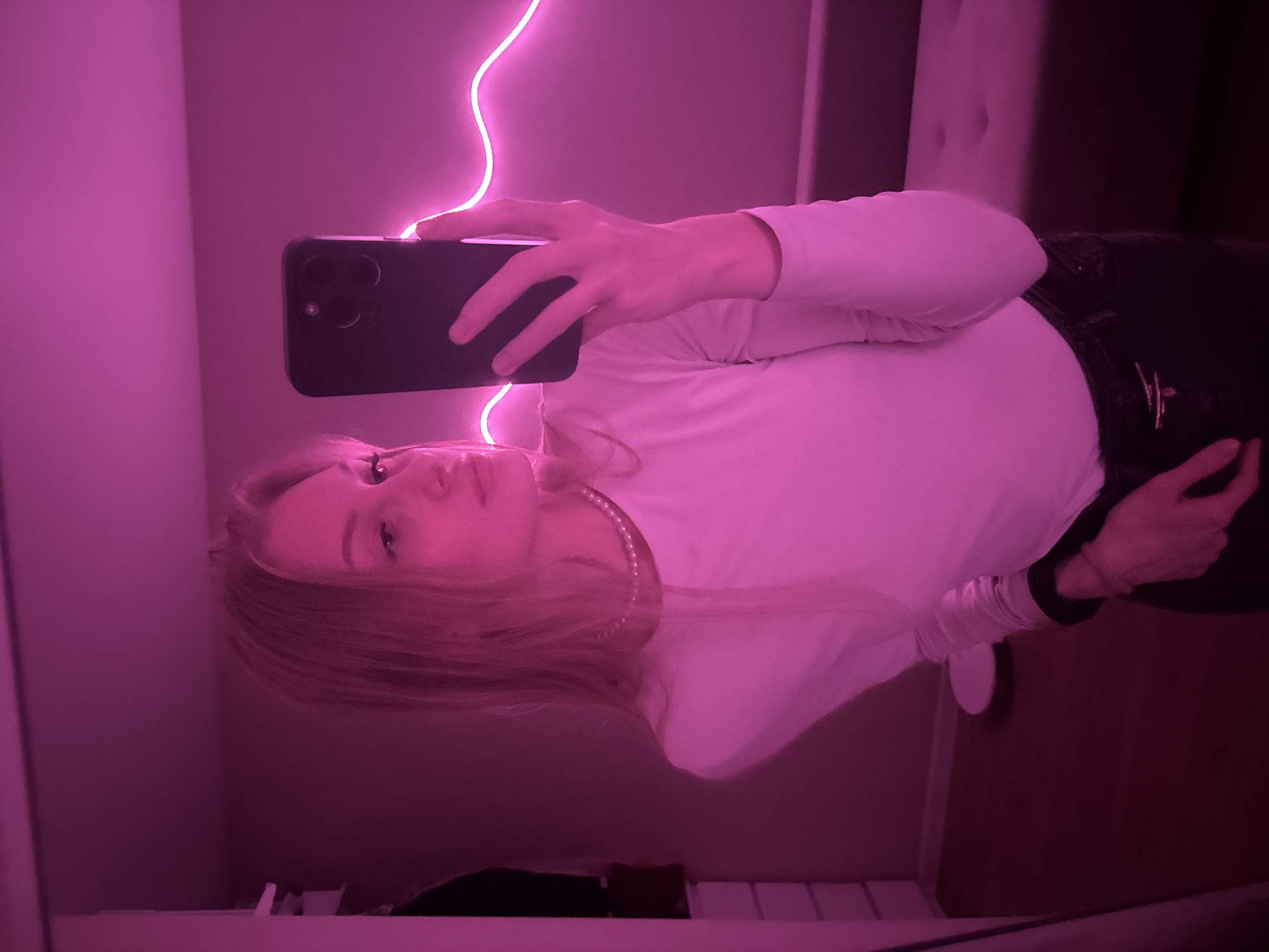 A mirror selfie captures a young person, appearing to be a female, holding a dark-colored smartphone with a prominent camera module in front of their face. The person has long, light-colored hair and is wearing a white long-sleeved top and a pearl necklace. Their expression is neutral as they look towards the camera's reflection. The background of the indoor scene is largely obscured by the strong, pervasive pink/magenta light emanating from a neon light strip that snakes vertically along the left side of the frame. Hints of a dark bed frame or headboard are visible in the upper right portion. The overall ambiance is dominated by this vibrant artificial lighting, creating a distinct, stylized atmosphere. There is no visible text in the image.