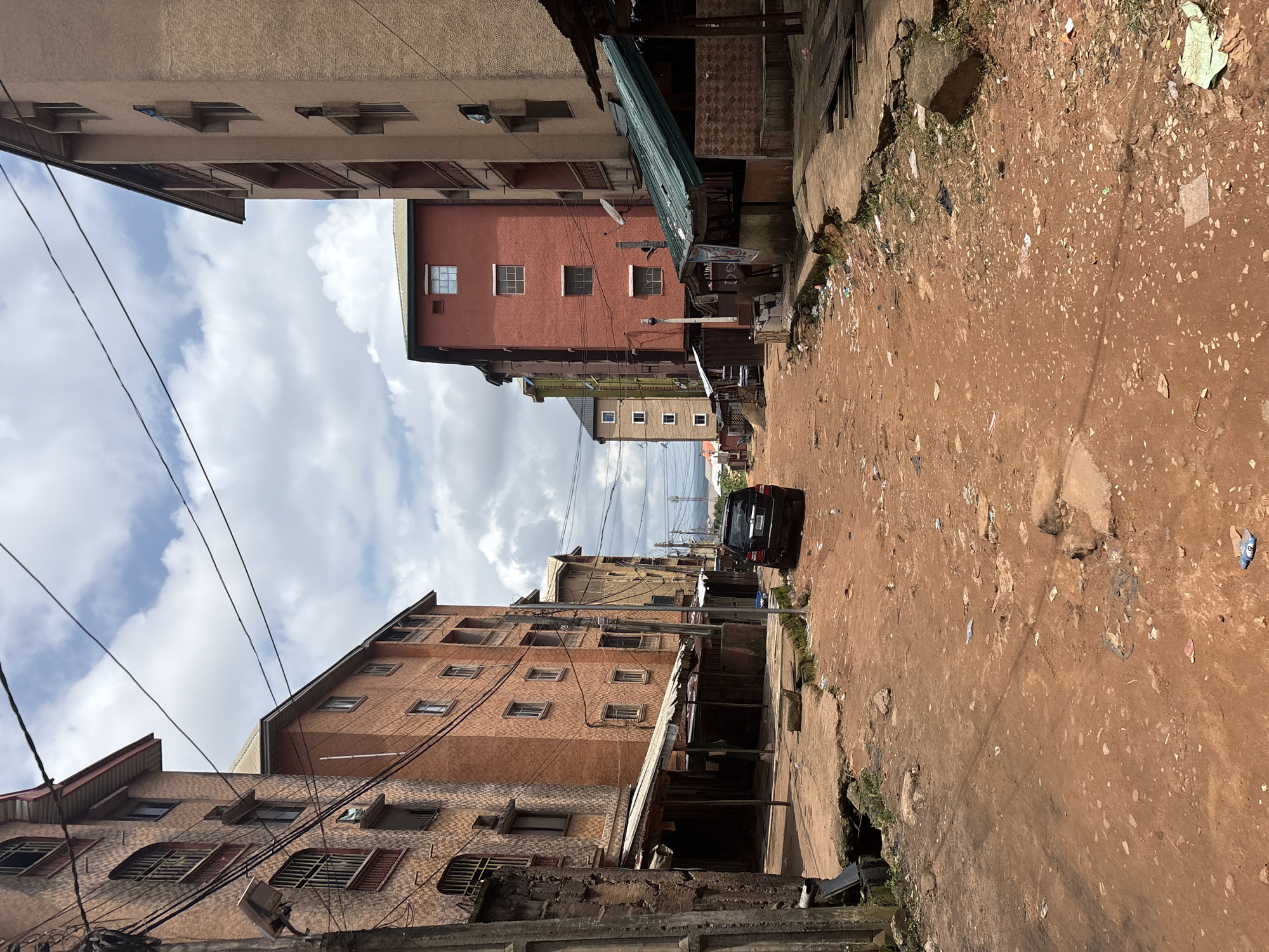 This is a street-level, medium-shot, eye-level photograph of a dirt road in Nkpor, Nigeria. The road is unpaved and appears to be made of red clay soil, with scattered debris, including plastic bags, paper, and small rocks. The road is flanked by multi-story apartment buildings on both sides. The buildings are constructed with concrete and brick, and many have balconies and windows with metal bars. Several small shops with corrugated metal roofs are set up along the sides of the road, in front of the apartment buildings.
On the left side of the road, an older, light-colored apartment building is visible, with a pole supporting electrical wires and a street light. A dark-colored SUV is parked on the right side of the road, facing away from the camera. The sky above is partly cloudy, with white clouds against a pale blue background. The scene suggests an urban or semi-urban environment with limited infrastructure.