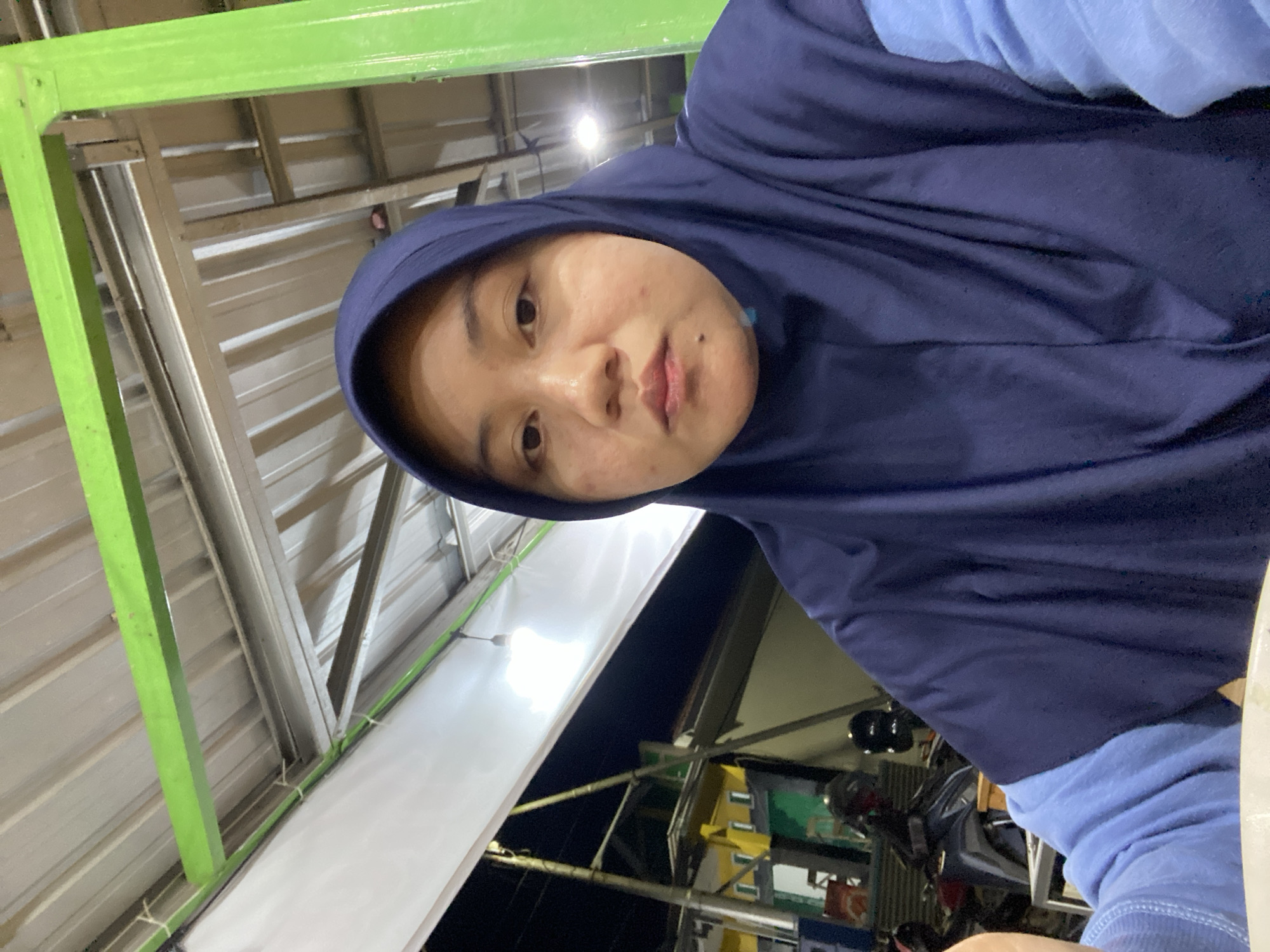 A young woman wearing a dark blue hijab and a light blue long-sleeved shirt is pictured in a selfie. She has fair skin and brown eyes, and she is looking directly at the camera. The background appears to be an outdoor covered area, possibly a food stall or market, with a corrugated metal roof supported by green metal beams. In the background, there are some motorcycles and buildings with yellow and green paint. The lighting suggests it is nighttime. This scene is likely located in the city of Banjarbaru, Indonesia, a region known for its warm climate and outdoor eateries.