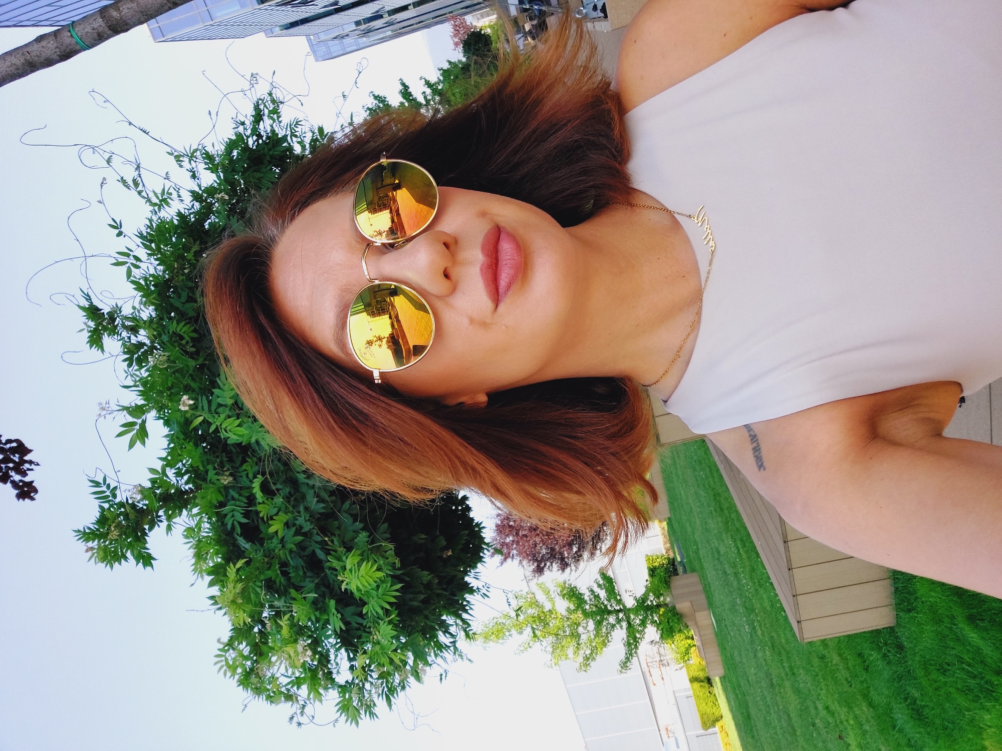 A woman with reddish-brown hair and gold-rimmed, circular mirrored sunglasses takes a selfie outdoors. She is wearing a light-colored, sleeveless top and a delicate gold necklace. A tattoo with Roman numerals is visible on her upper left arm. The background shows a lush green lawn with wooden benches, a variety of trees, and modern buildings, suggesting an urban park or courtyard setting in Sofia, Bulgaria. The sky is clear and bright.