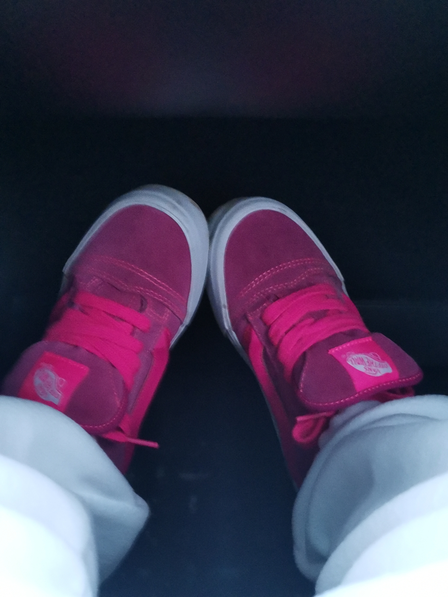 The image shows a close-up, top-down view of a person's feet wearing bright pink high-top Vans sneakers. The sneakers have white laces and white soles. The person is wearing white sweatpants or joggers. The background is dark and out of focus, suggesting an indoor or dimly lit environment. The scene does not provide any information about the location in the city of 29-Y Km, Russia, as no contextual elements or landmarks are visible.