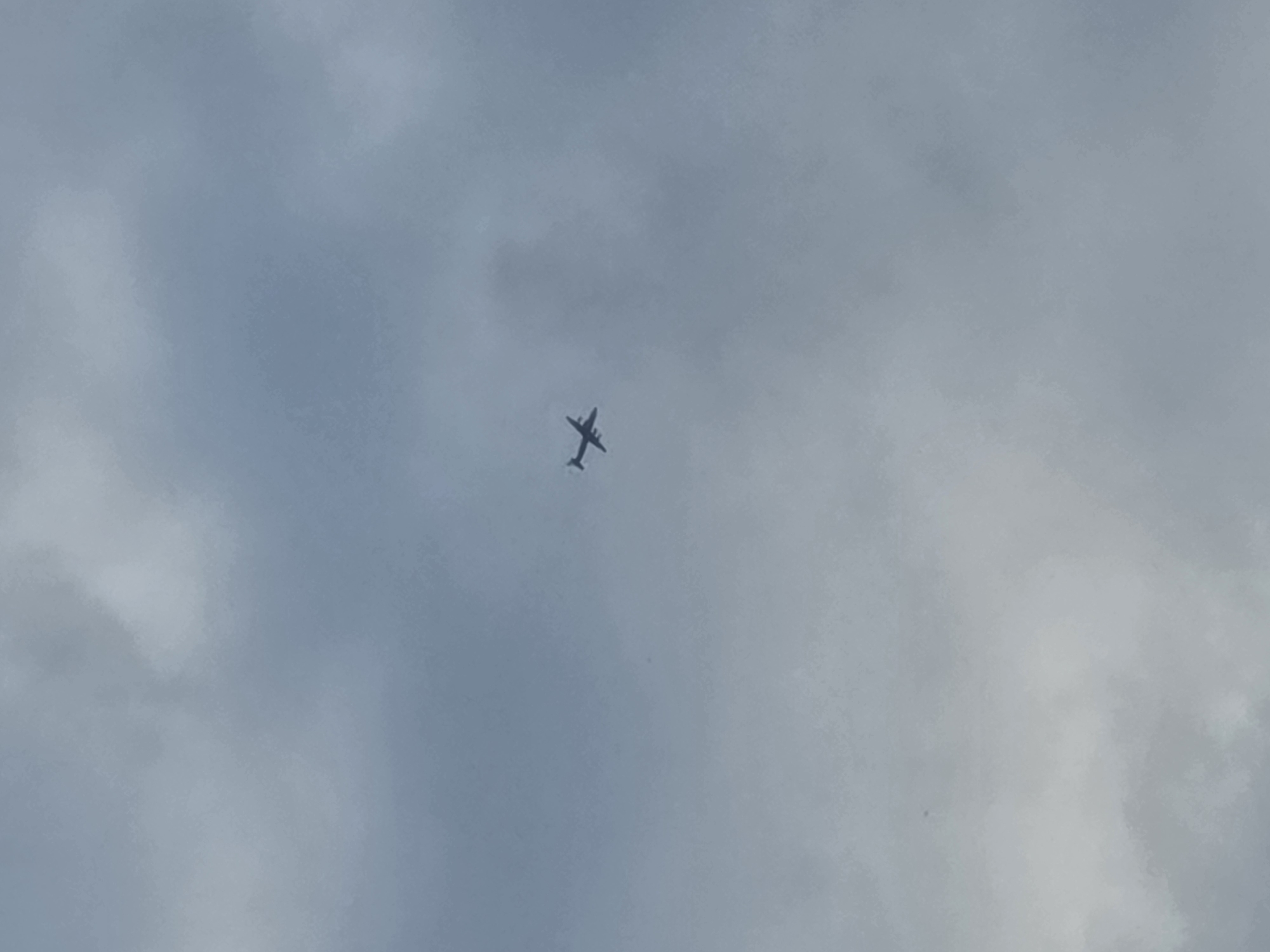 A single airplane is visible in the sky, silhouetted against a cloudy backdrop. The plane is dark and appears to be in flight. The sky is overcast with varying shades of gray clouds. The image does not provide any specific landmarks or context to pinpoint a location in Frolovo, Russia.