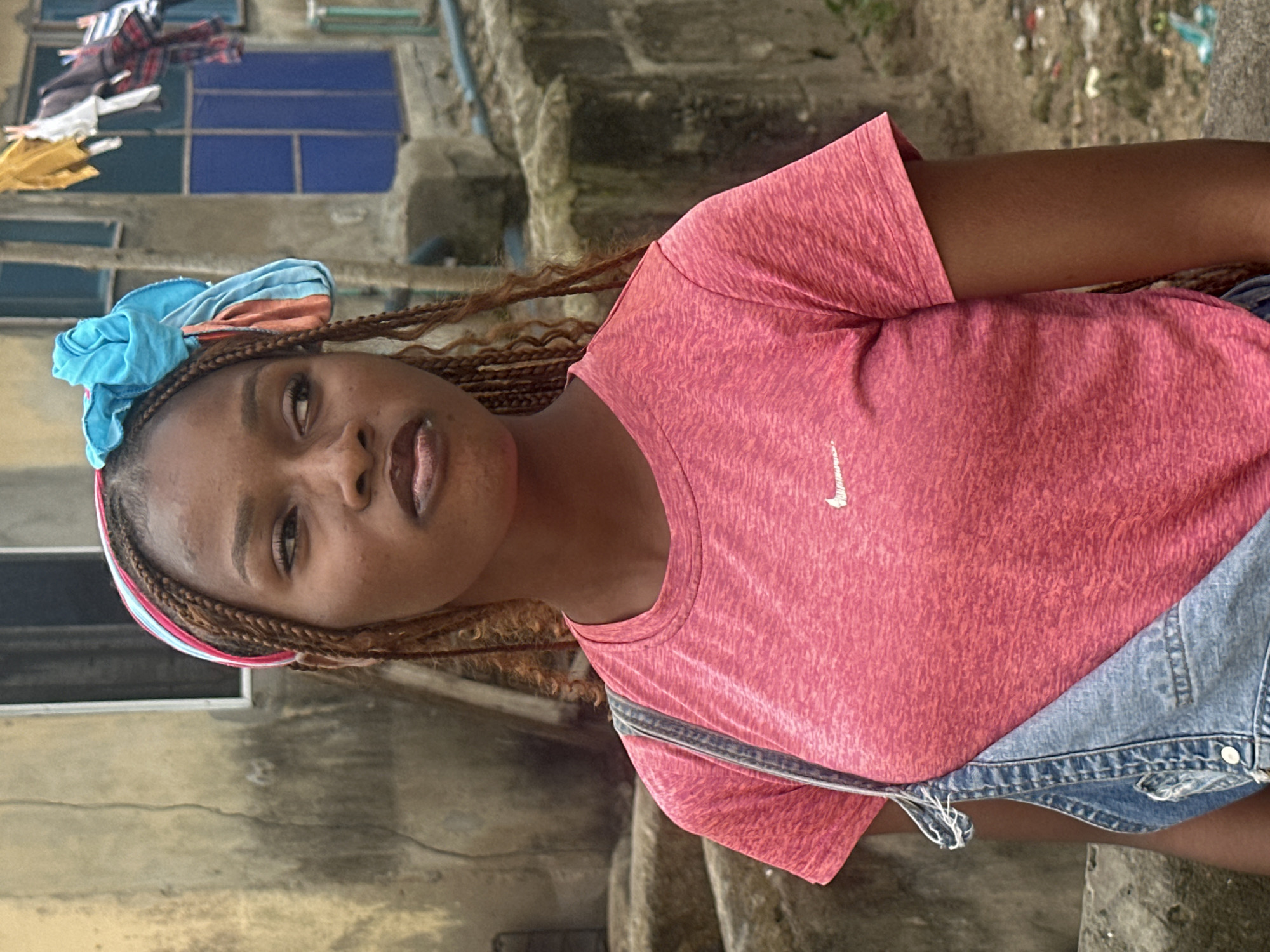 A medium shot captures a young Black woman standing outdoors, looking to her left. She has dark skin, braided hair, and is wearing a pink t-shirt with a white logo, and denim overalls. A colorful headscarf, predominantly blue with hints of orange and pink, is tied around her head, with some fabric cascading down the side of her head. Her hair is braided and hangs down her back. The background appears to be an urban environment in Badagry, Nigeria, with a weathered building wall behind her. Laundry is hanging on a line in front of a blue-framed window in the distance. A concrete structure with stairs is visible to the left, and a low concrete wall is in the foreground. The ground is dirt and gravel.