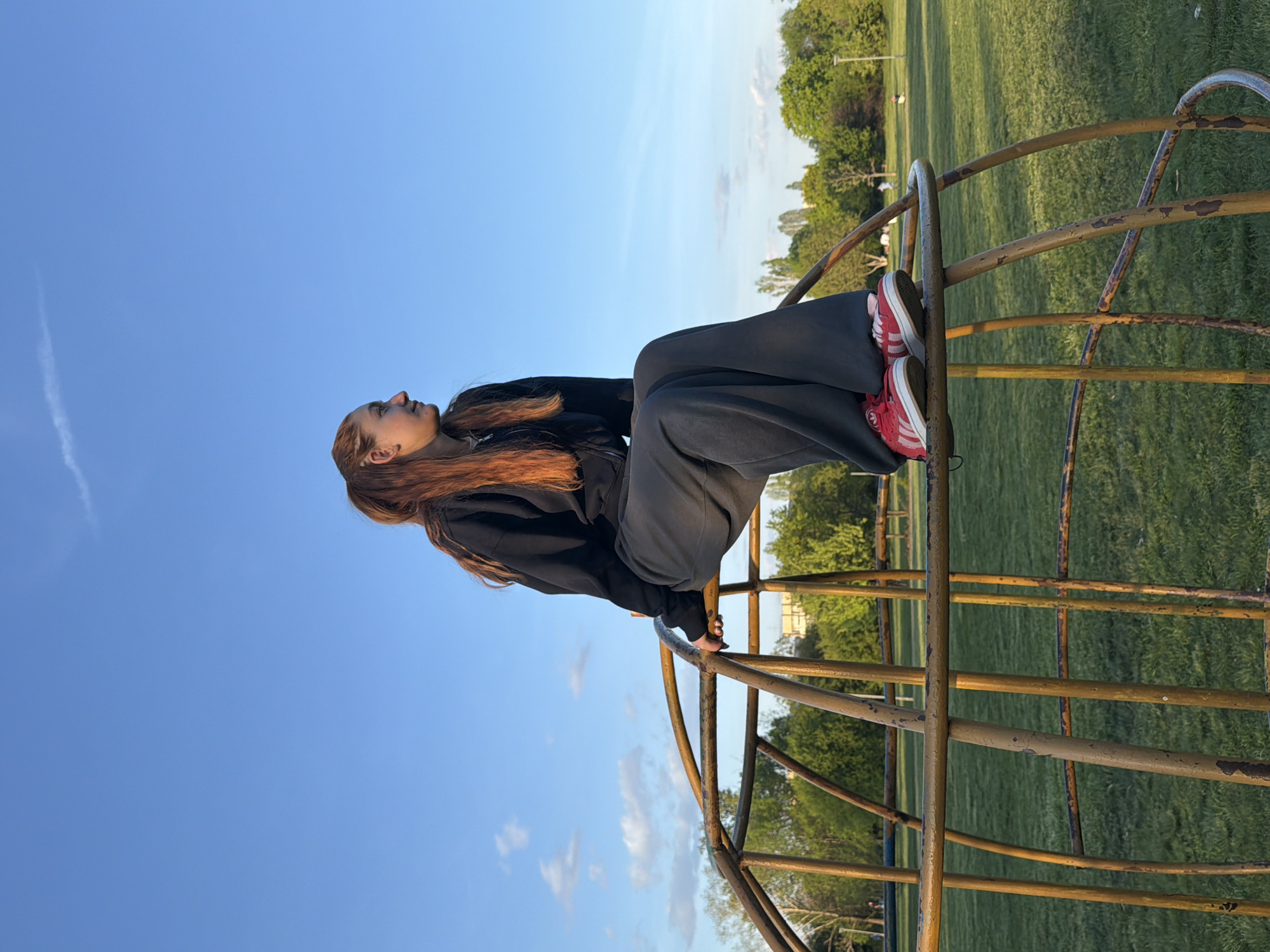 A young person is depicted sitting on an old, yellow, metal playground structure in an outdoor park setting in Sofia, Bulgaria. The image is oriented vertically, showing the person on the right side looking upwards.
The subject has long, reddish-brown hair and is wearing a black long-sleeved top or hoodie, and dark, wide-legged pants. On their feet are distinctive red sneakers with white stripes, possibly an Adidas model. They are positioned with their knees bent and are looking towards the upper left of the frame, with a calm and contemplative expression. Their hands are resting near the metal bars.
The playground structure they are sitting on appears to be a large, spherical or domed climbing frame, showing signs of age with chipped yellow paint and some rust.
The background features a vast, clear blue sky occupying a significant portion of the image, with a faint white contrail visible towards the bottom left. Below the horizon line, a lush green grassy area extends into the distance, dotted with numerous trees under a bright, sunny sky. The lighting suggests it is daytime, possibly late afternoon, given the warm glow on the person's hair and face. The overall atmosphere is peaceful and open. There is no visible text in the image.
