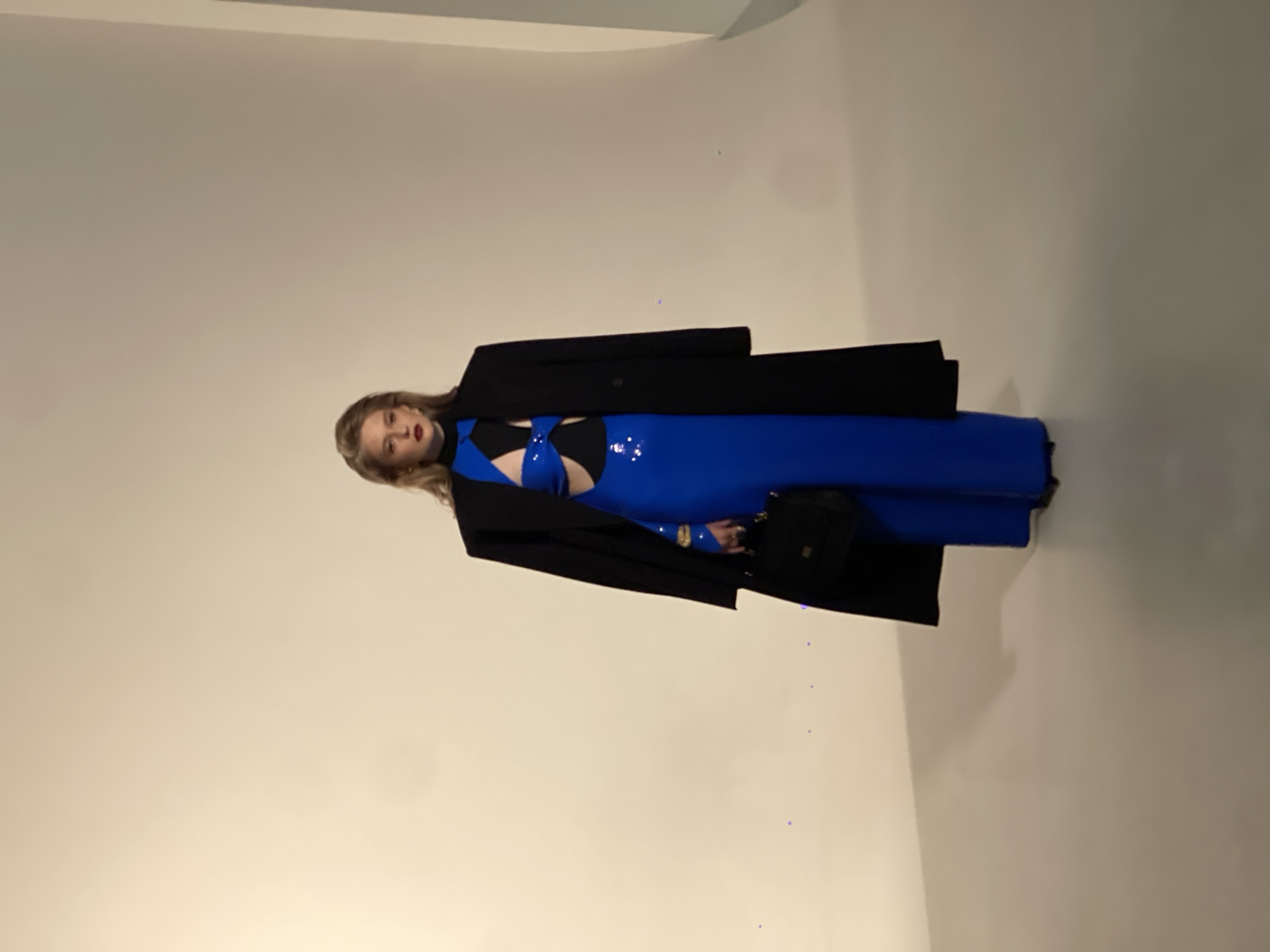 A woman stands in a studio against a plain backdrop. She is wearing a long, bright blue dress with cutouts at the waist and chest, and a black jacket draped over her shoulders. She has dark hair, is wearing red lipstick, and is carrying a black handbag. The floor is light gray. The image appears to be a fashion photograph.