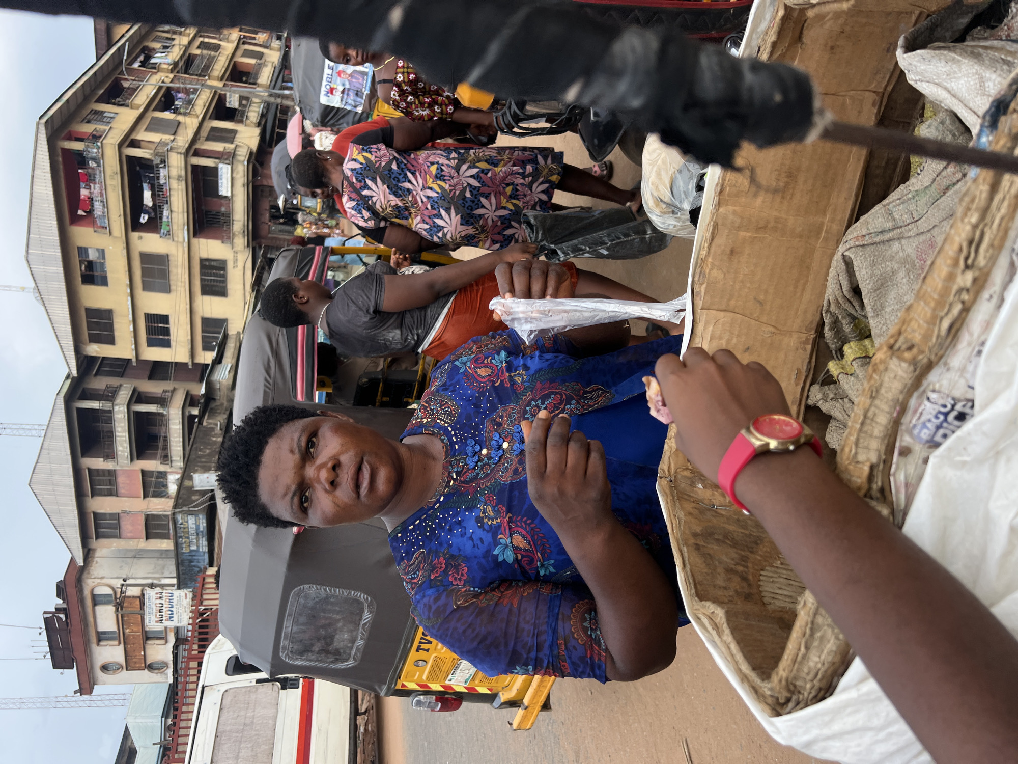 In Nkpor, Nigeria, a woman with dark, short, curly hair and a serious expression is captured at eye level. She is wearing a vibrant blue dress adorned with a colorful floral and beaded pattern. Her right hand is extended, holding a small, light-colored object, possibly food, and a clear plastic bag. In the foreground, a person's arm with a red watch is reaching into a large, worn cardboard box filled with sacks. The background reveals a bustling street scene with a yellow and grey tricycle or auto-rickshaw partially visible. Several multi-story buildings with balconies and laundry hanging out are also present, suggesting a commercial or residential area. The overall lighting indicates an overcast day, typical of the Nigerian climate.