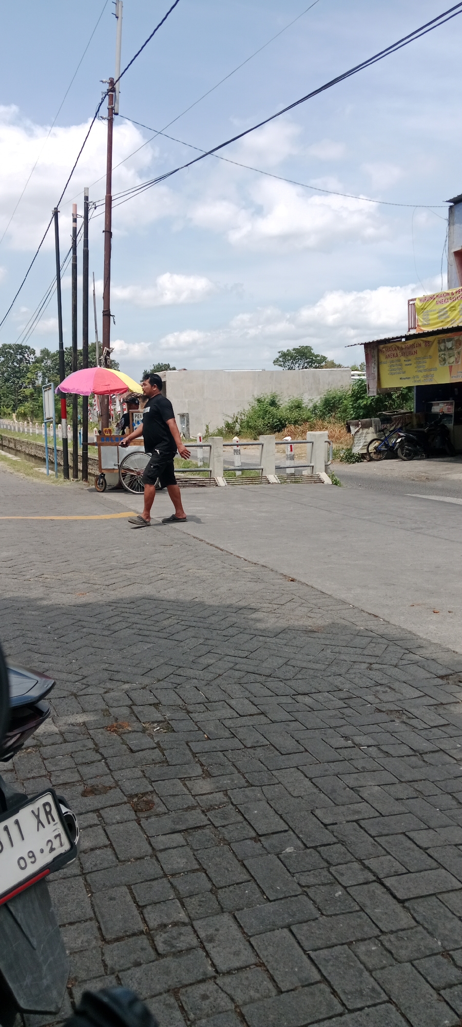 In Maguwoharjo, Indonesia, a man in a black t-shirt and shorts walks across a gray paved road. He is wearing flip-flops. To his left, a colorful umbrella adorns a food cart. In the background, a building with a yellow sign and several motorcycles are visible. A motorcycle license plate, reading "B11 XR 09.27", is partially visible in the foreground. Overhead, telephone wires crisscross a blue sky with scattered clouds.