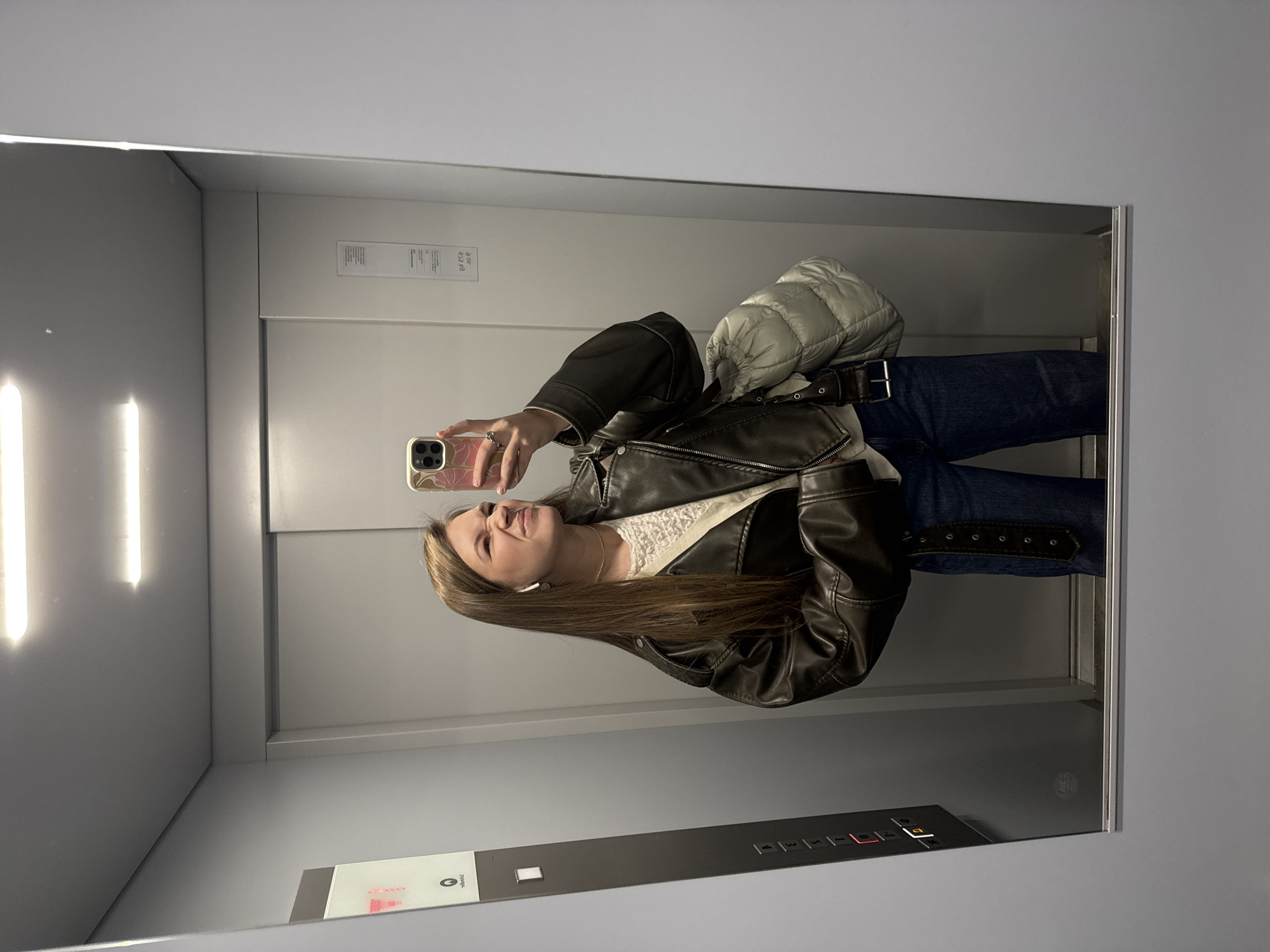 A young woman is captured in a mirror selfie inside an elevator. She has long, straight brown hair and is wearing a dark brown leather jacket over a white, textured top, and blue jeans with a dark belt. A light-colored puffer jacket or bag is draped over her left shoulder. White wireless earbuds are visible in her ears. She holds a smartphone with a floral-patterned case in her right hand, positioned to take the picture, and displays a slight smile.
The setting is an elevator with reflective walls. Bright, rectangular light panels illuminate the space, visible on what appears to be the left wall. The elevator control panel is visible at the bottom of the reflection, featuring numbered buttons from "1" to "6", along with symbols for door close, door open, a bell, and a phone. The "1" button is lit red, and the "6" button glows orange. Above these buttons, a circular symbol is present. Mirrored text, likely a location or brand name, appears on the control panel, which reads "Vozdovac" when viewed correctly. Additionally, faint, mirrored instructional text or capacity information is visible on a panel on the right side of the elevator, reflecting as if on the ceiling or upper part of the wall, with some numbers like "80 kg" potentially discernible. The overall mood is casual and relaxed.