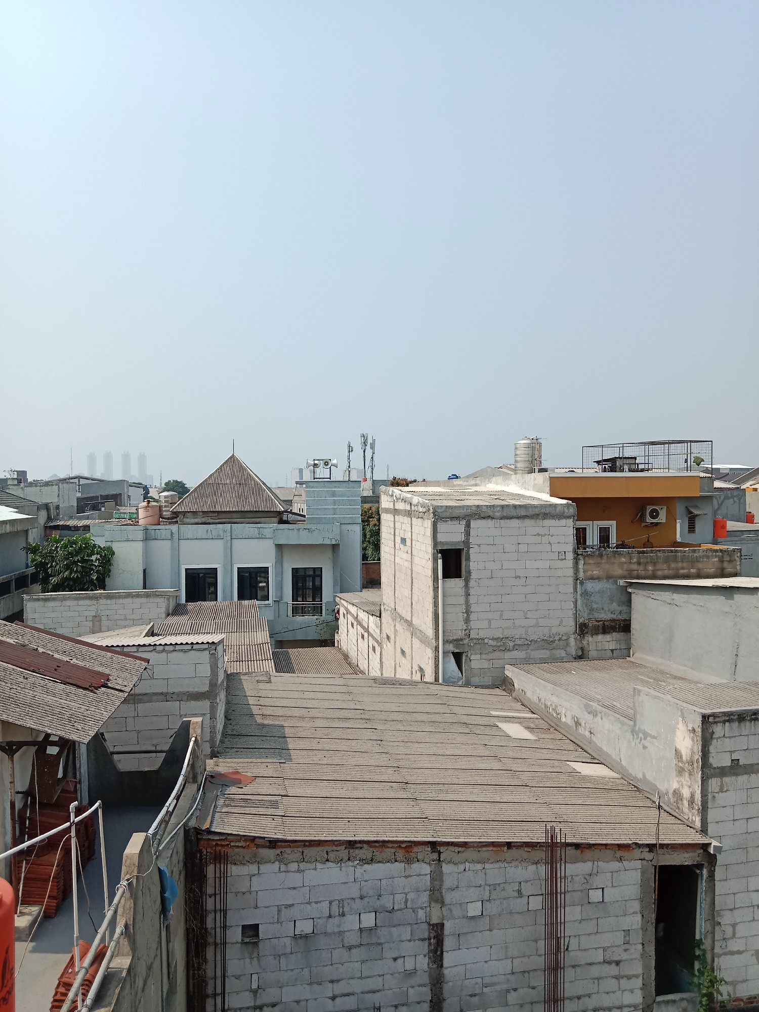 This is a daytime, eye-level, medium shot captured in West Jakarta, Indonesia, featuring a dense collection of urban rooftops. The scene is dominated by various buildings constructed from concrete blocks and corrugated metal roofing. In the foreground, several rooftops are visible, some with exposed metal rebar, suggesting ongoing construction or renovation. A stack of orange-colored tiles is visible on the left side of the image.
Further back, a light gray building with a distinct pyramidal roof stands out. This building has several windows with black frames. Adjacent to it, a light orange building features an air conditioning unit and a caged rooftop area. The middle ground shows more residential or commercial structures, some appearing unfinished. In the distance, several tall, modern buildings are silhouetted against the hazy sky, indicating a more developed urban area. The sky is clear and pale blue, suggesting a bright, possibly humid day. No people are visible in the image. The overall impression is one of a developing urban landscape with a mix of older and newer construction.