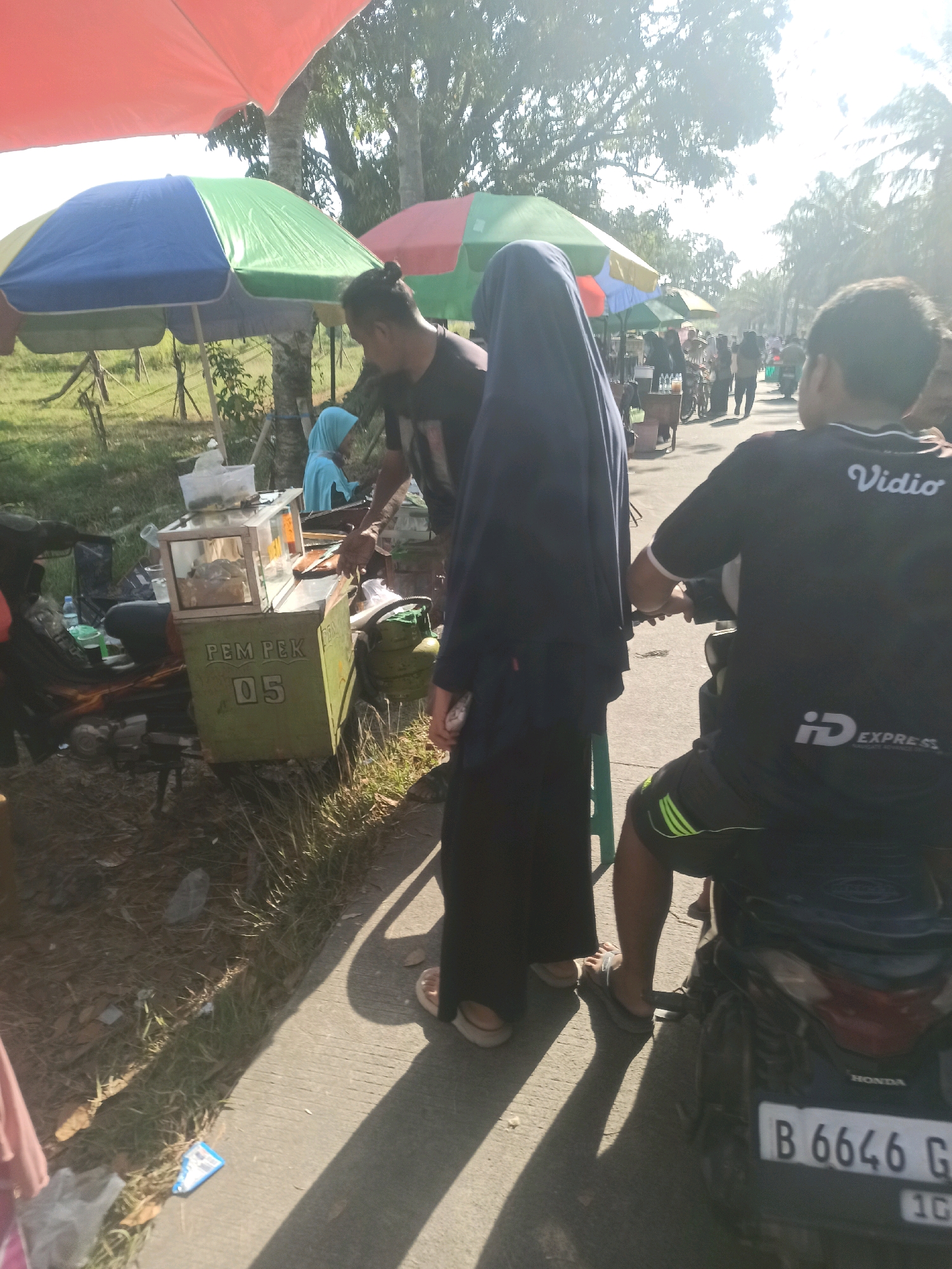 In Telagamurni, Indonesia, a street scene unfolds under a sunny sky with dappled sunlight filtering through trees. A food cart, painted green and labeled "PEM PEK 05," is situated on the right side of a paved road. On the cart, a glass display case holds food items, with a gas cylinder positioned behind it. Two individuals are visible near the cart. One, wearing a dark shirt and a blue headscarf, appears to be interacting with the cart. Another person, clad in a dark blue abaya, stands to the left of the food cart, facing away from the camera. To the right of the scene, a person wearing a black t-shirt with "Vidio" and "ID EXPRESS" logos, and striped shorts, is seated on a black motorcycle. The motorcycle has a license plate that reads "B 6646 G". In the background, other individuals and motorcycles are present, suggesting a bustling market or roadside gathering. Colorful umbrellas are set up, providing shade over some areas of the scene. The ground is a mix of dirt, grass, and paved road, with scattered debris.