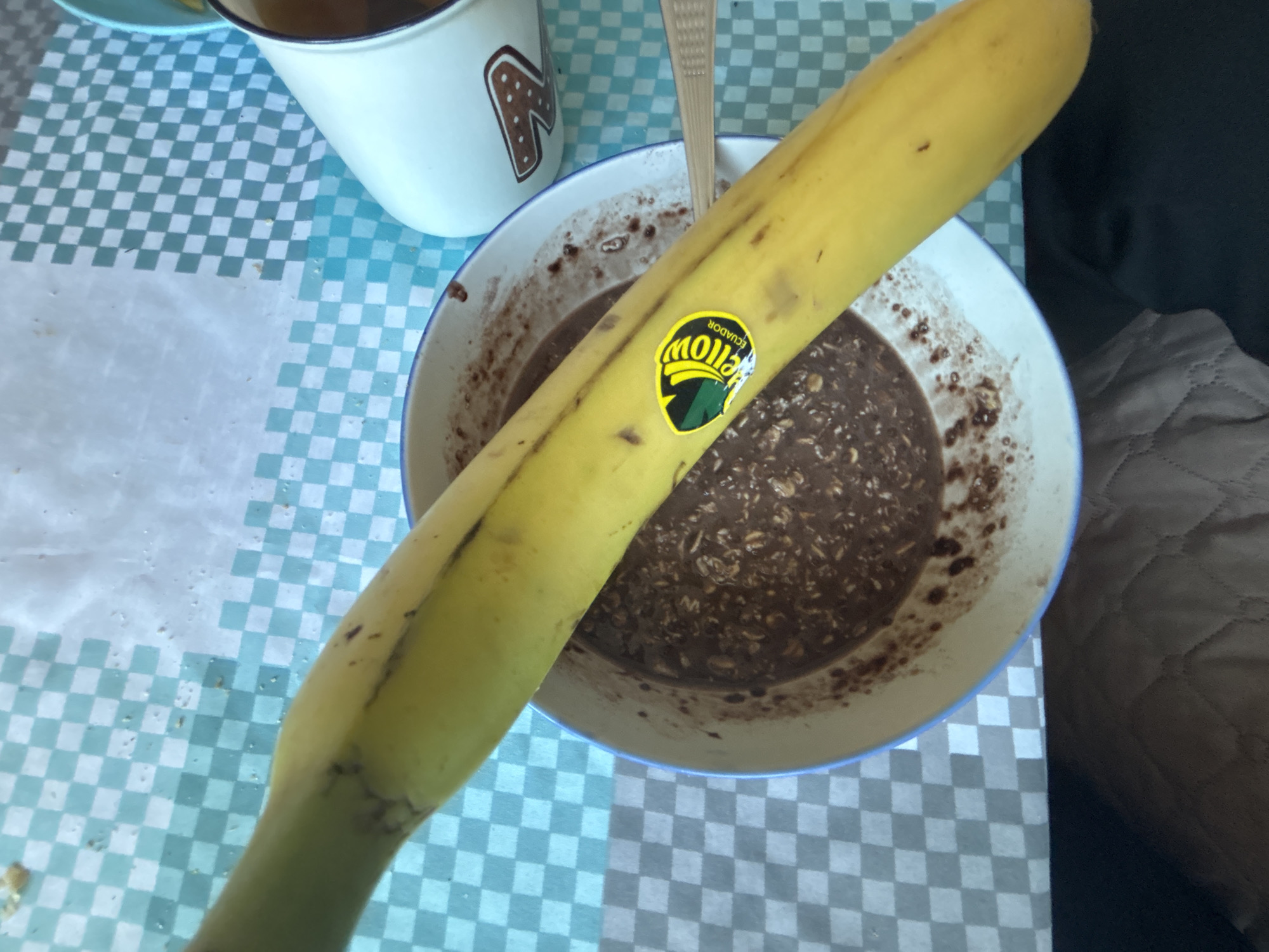The image shows a table with a blue and white checkered tablecloth. A white mug with a brown letter "N" design and a bowl containing a dark, possibly chocolatey mixture with oats are present. A banana with a "Yellow Ecuador" sticker is placed across the bowl. A spoon is in the bowl. A portion of a brown couch is visible in the background on the right. There is no information in the image to suggest a location in the city of Zlatograd, Bulgaria.