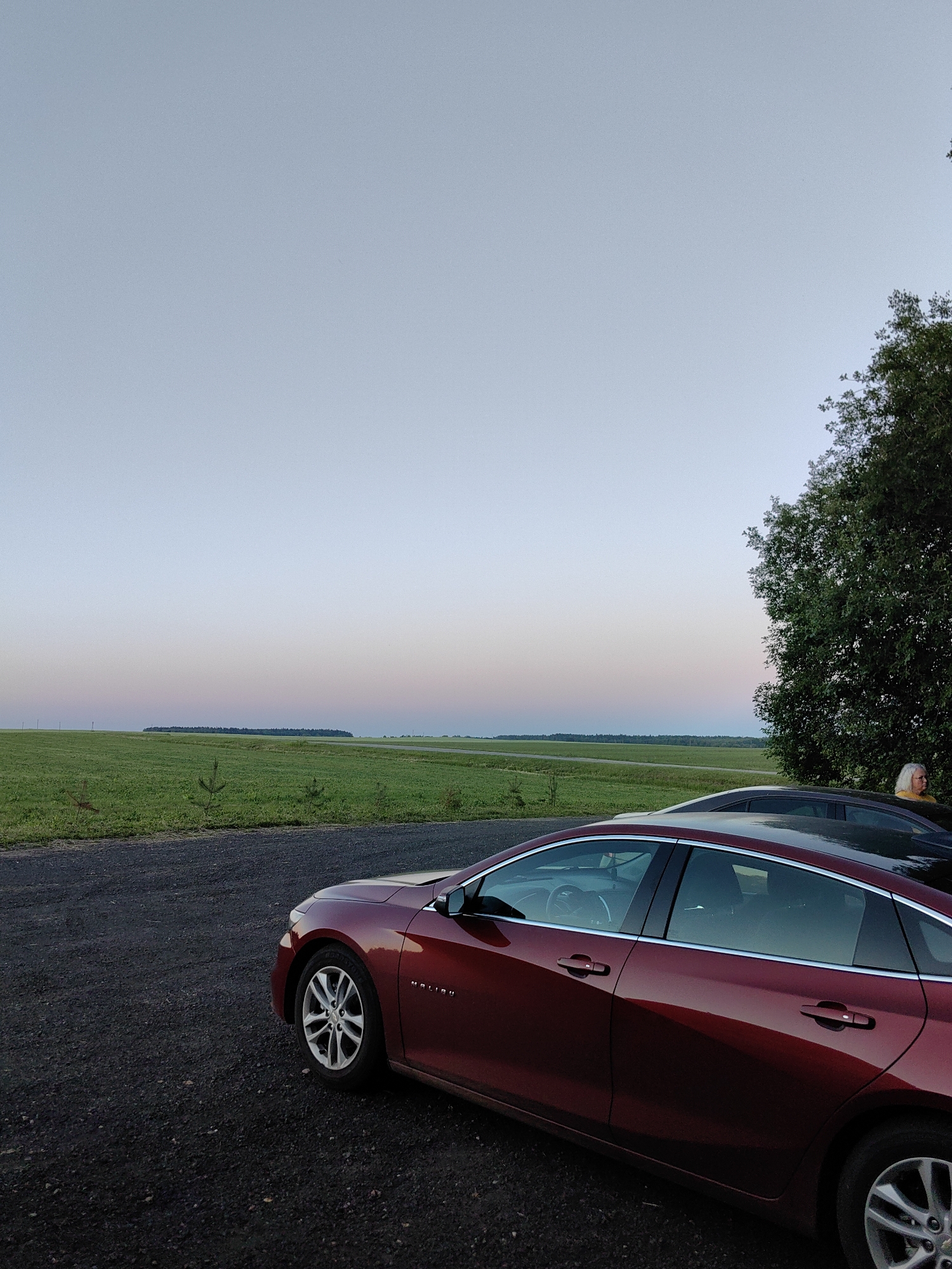 The image shows a red Chevrolet Malibu parked on a gravel surface, with a dark-colored car partially visible behind it. A woman with white hair, wearing a yellow top, is standing near the dark-colored car, looking to the right. The cars are facing left, with the red car in the foreground. Behind the cars, there is a vast green field that stretches to the horizon, with a line of trees in the distance and a subtle gradient of pale blue, pink, and yellow in the sky, suggesting it is either dawn or dusk. The scene is set in an outdoor environment, likely in the countryside, given the expansive field and the absence of urban structures. The overall impression is one of a quiet, open landscape, with the cars and the woman as the primary points of human presence. The location is identified as Дзямідавіцкі сельскі Савет, Belarus.