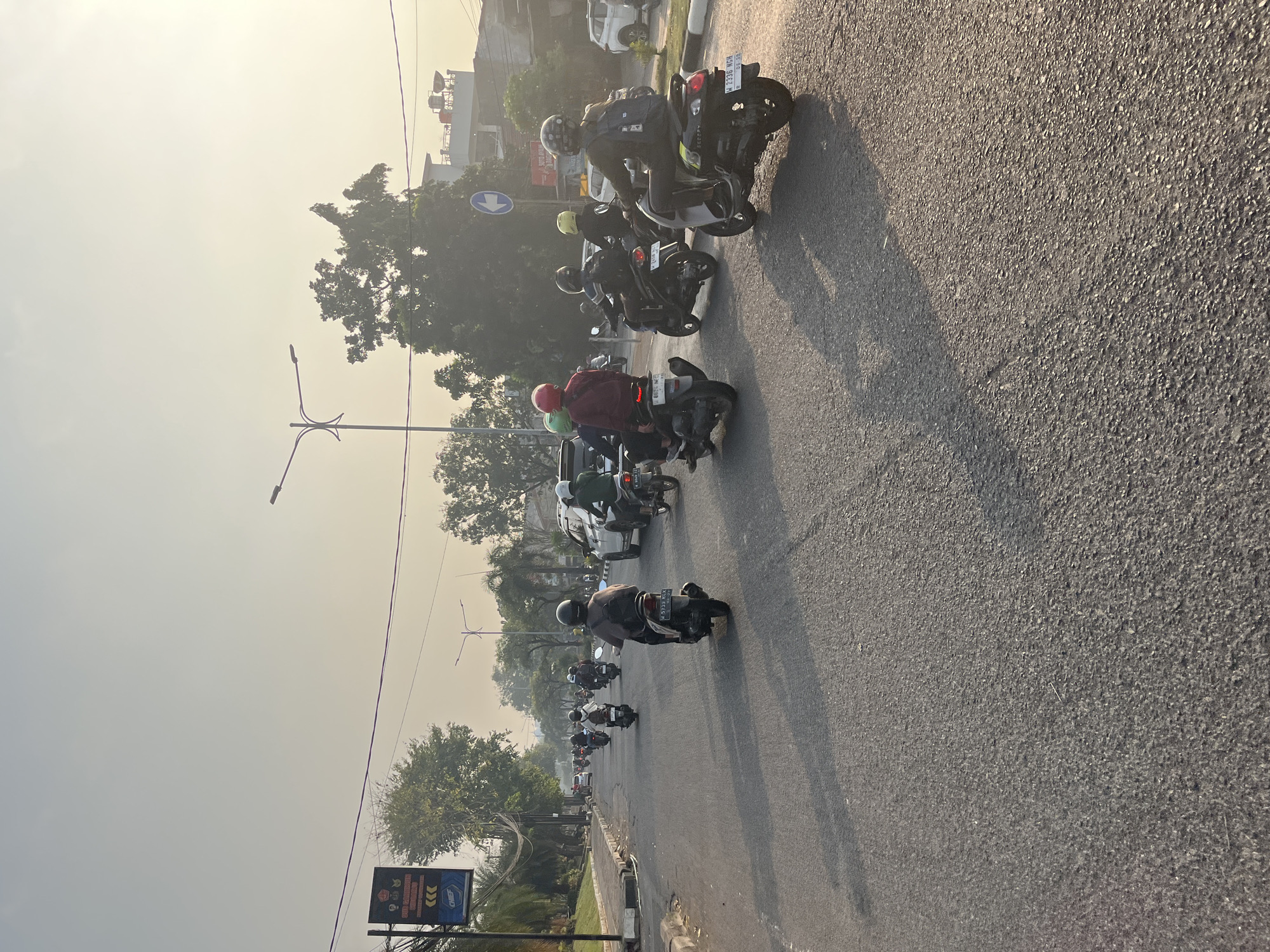 In Tenggulunan, Indonesia, on a hazy, sunlit morning, a procession of motorcycles navigates a paved road. The asphalt is marked with faint lane divisions and softened by long shadows cast from the left. The street is lined with a concrete curb and a raised median, bordered by a grassy verge and trees. A signpost on the left displays various symbols and text, including "SUB KANTOR PENGURUSAN SURABAYA." In the distance, streetlights and electrical wires crisscross the sky. The motorcyclists, clad in helmets of various colors, are the primary subjects, each on their own scooter or motorcycle, traveling in the same direction. Some wear backpacks, and a few are accompanied by passengers. A white car and a white SUV are also visible, interspersed within the motorcycle traffic. The overall atmosphere is one of daily commuting in a moderately urban environment under a soft, overcast sky.