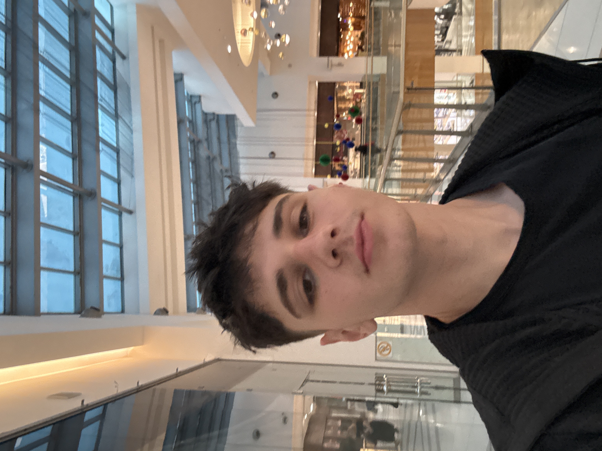 A young man is featured in a close-up selfie taken indoors, likely within a modern shopping mall or large commercial building in Varna, Bulgaria. He has short, dark hair and a neutral expression, looking directly at the camera. He is wearing a black top or jacket.
The background reveals a multi-story interior with contemporary architectural elements. To the left, large, multi-paned windows allow bright natural light into the space. The right and deeper background show several levels of the building, featuring glass railings and what appear to be escalators or stairs. Overhead, spherical decorative light fixtures hang from a high ceiling. In the mid-ground, colorful spherical decorations in shades of red, blue, green, and yellow are suspended, adding a festive touch. Reflections of the interior are visible on the polished floor surface. Faintly visible in the far background is a circular yellow sign, possibly indicating a prohibition or warning. The overall lighting suggests it is daytime.