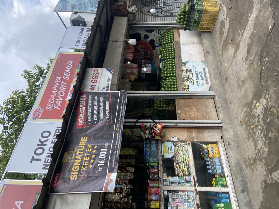 The scene is a small storefront in Lembang, Indonesia, located at the coordinates -6.817 latitude and 107.613 longitude. The store is a simple structure with a wooden frame and a corrugated metal roof. The storefront is covered in signs in Indonesian and English. One sign advertises "Signature Kretek" cigarettes for 16,000 Indonesian Rupiah. Another sign advertises "Revolusi Kretek," which appears to be a brand of cigarettes. The store window is filled with packaged goods, including drinks, snacks, and tobacco products. The store is open and there is a small amount of product displayed inside and outside the store. There are stacks of avocados in the corner, and a sign in Indonesian says "DISINI BISA BIKIN JUS ALPUKAT!!" which translates to "Here you can make avocado juice!". There is a stack of green avocados behind the store window. The storefront is located on a busy street with a concrete sidewalk and asphalt street. There are other buildings and structures visible in the background. The weather is overcast and grey. There are no people in the photo.