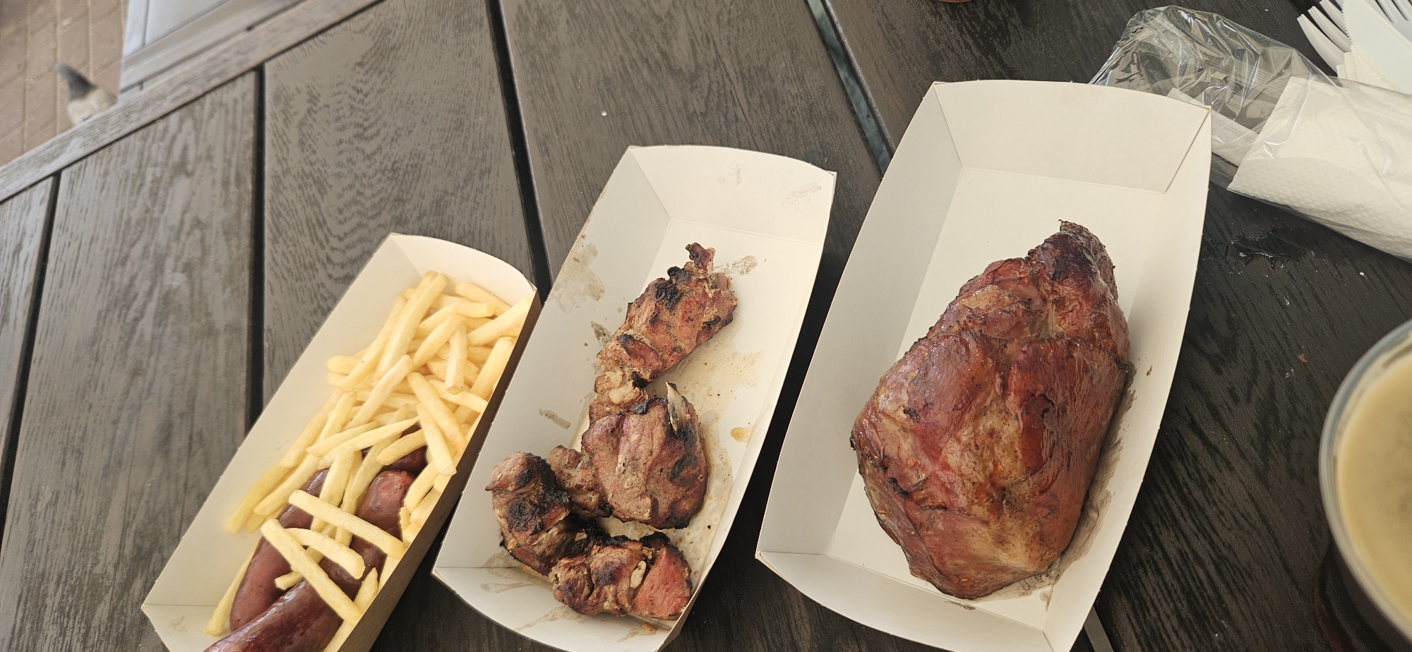 A top-down medium shot shows three white paper trays of food arranged on a dark wooden table. The first tray, at the top of the frame, contains several cooked sausages and a pile of french fries. The second tray, in the middle of the frame, holds pieces of grilled meat. The third tray, at the bottom of the frame, contains a large piece of roasted meat. The table surface has a visible wood grain pattern. In the upper right corner, a blurred object resembling a pigeon is visible on a brick surface. There are also some plastic utensils and a napkin visible on the table. The scene appears to be outdoors, possibly at a restaurant or picnic area. There is no indication of a specific location in Hrodna, Belarus.