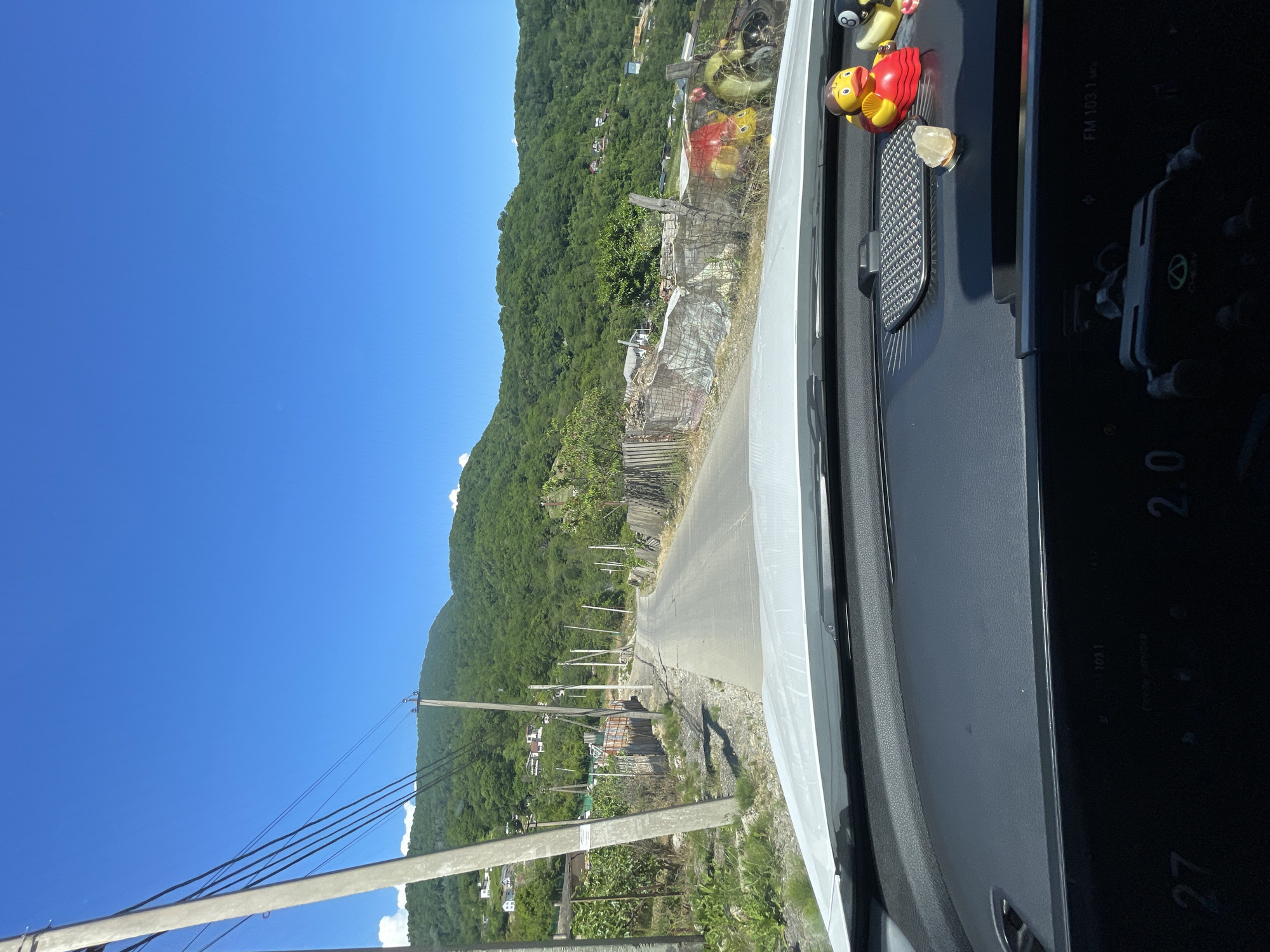 The image shows a view from inside a vehicle, looking out onto a paved road leading through a hilly, green landscape under a clear blue sky. The road is lined with vegetation, utility poles, and fences made of wood and corrugated metal. Some houses are visible on the slopes of the hills. In the foreground, on the dashboard of the vehicle, there are several decorative items, including two rubber ducks and what appears to be a billiard ball. The car's dashboard displays some digital information, including the numbers "27" and "2.0" and "FM 103.1". The location is identified as Verkhniy Yurt, Russia.