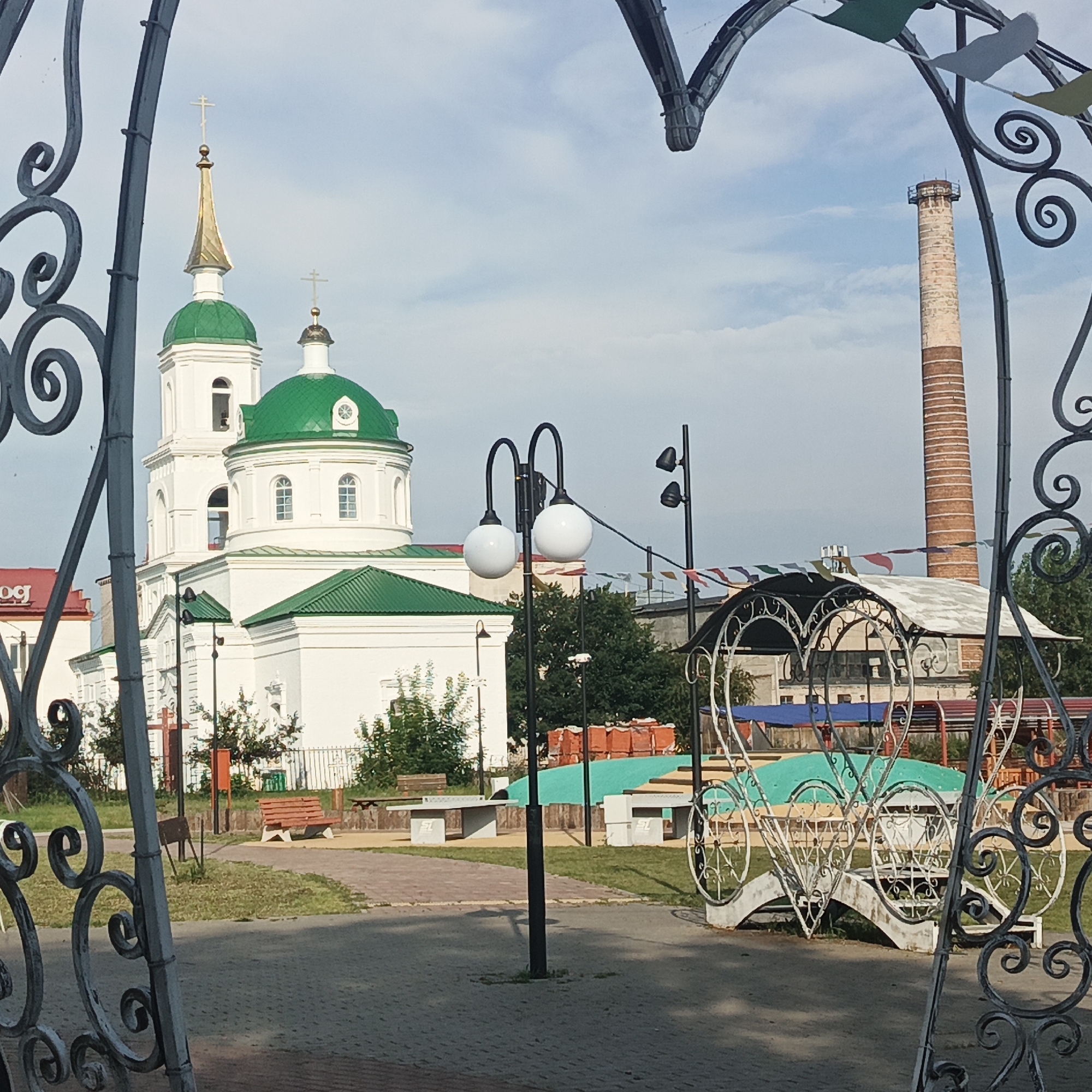 The image captures an outdoor scene in Kambarka, Russia, featuring a white church with green domes and a tall brick chimney in the background. In the foreground, decorative metalwork arches frame the view. The church, which appears to be an Orthodox church due to the crosses on its spires, has a prominent bell tower and several domes.
Adjacent to the church, there are pathways, grassy areas, benches, and what looks like a park or recreational area. These include ping pong tables, a brightly colored surface that might be a play area, and a white decorative gazebo or seating area with intricate metalwork. Benches are scattered throughout the area, and some trees and bushes are visible.
The brick chimney is a distinctive industrial element in the landscape, suggesting the presence of industry in the vicinity. The sky is partly cloudy, indicating daytime. The overall impression is a mix of historical architecture, recreational space, and industrial elements, characteristic of a town like Kambarka.