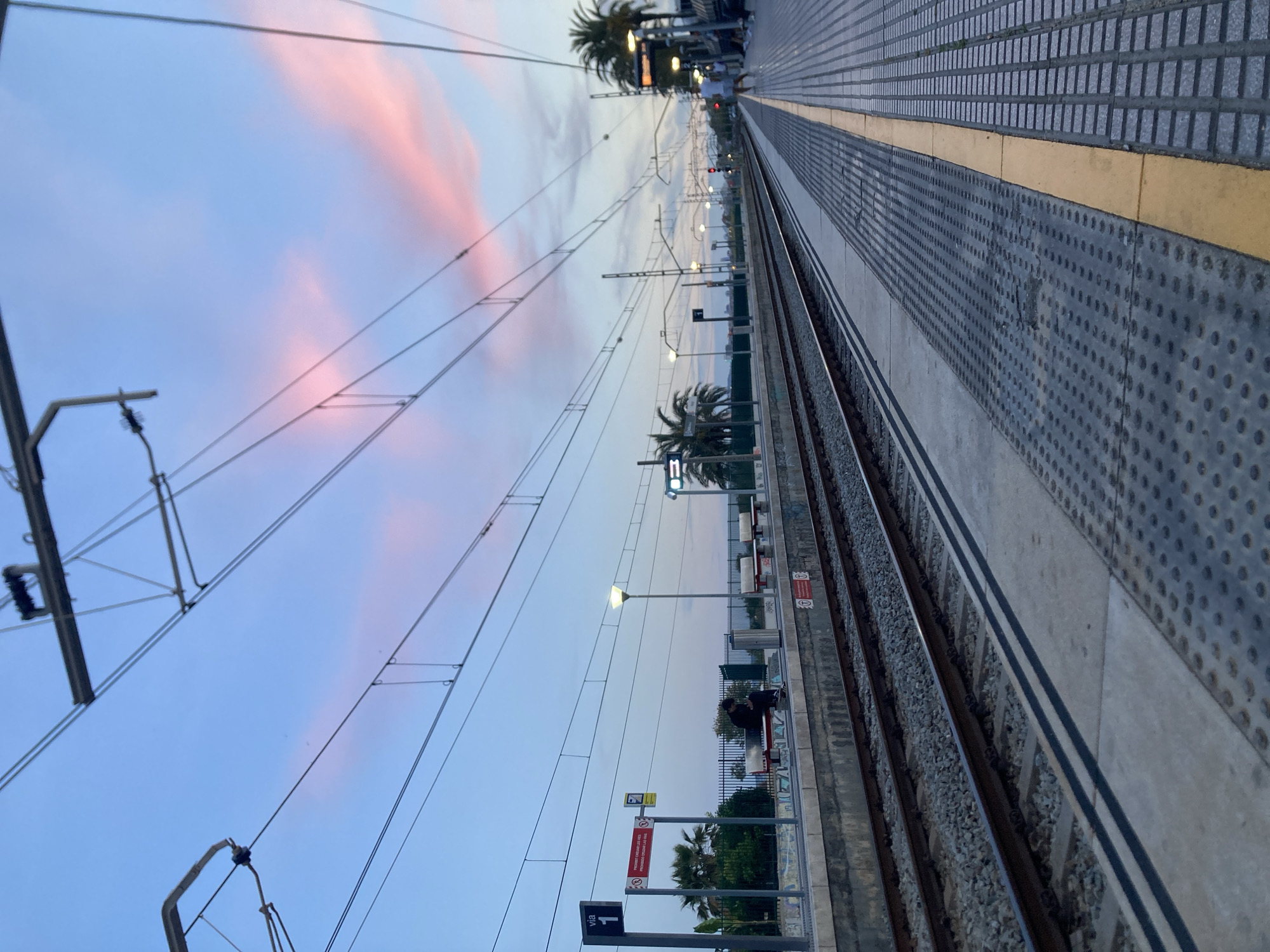 The image shows a train platform in Montgat, Spain, at dusk. The platform is constructed of patterned concrete with a yellow safety strip. Railroad tracks run parallel to the platform. Overhead electrical lines are visible against a sky with pink-tinged clouds. Palm trees are scattered along the view from the platform. A few people are visible sitting on benches along the platform. Signage is posted, including a "Via 1" sign and a warning sign in Spanish and Catalan. Streetlights illuminate the area.