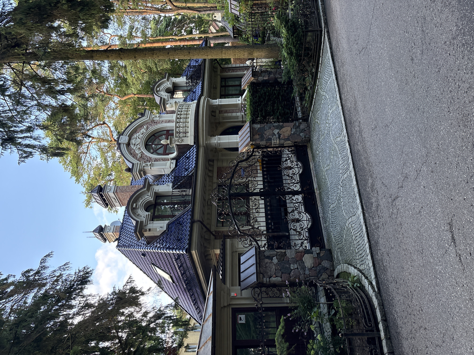 The image shows a large, ornate house with a dark blue tiled roof, surrounded by trees. The house has multiple stories, dormer windows, and decorative architectural elements. A large, black wrought-iron gate stands in front of the house, partially obscuring the driveway, which is paved with cobblestones. Stone pillars topped with metal accents support the gate. A security camera is visible on the left side of the gate. The setting appears to be a residential area, likely in a town or city, with other houses and trees visible in the background. The sky is partly cloudy. The image is taken from a street-level perspective.
