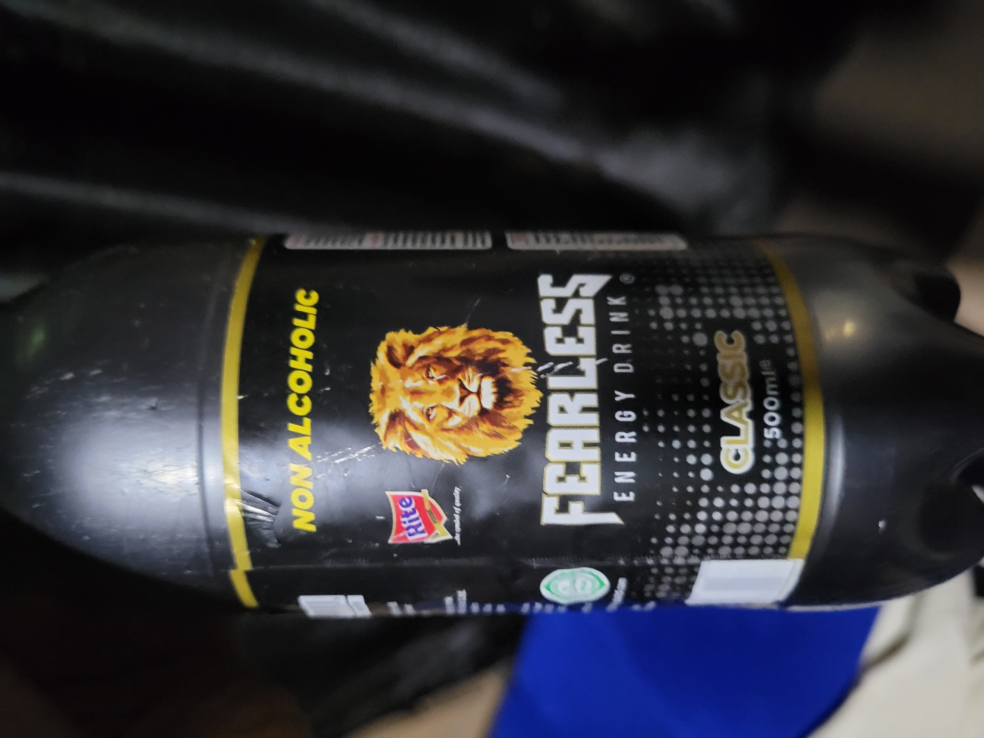 A close-up, eye-level, indoor shot shows a black plastic bottle of "FEARLESS ENERGY DRINK" with a yellow and white label. The label features a lion's head, the text "NON ALCOHOLIC," "FEARLESS ENERGY DRINK," and "CLASSIC 500ml". A red and white "Rite Foods" logo is also visible. The bottle is positioned vertically and takes up most of the frame, with the background out of focus, showing dark fabric. No people or specific location details are discernible.