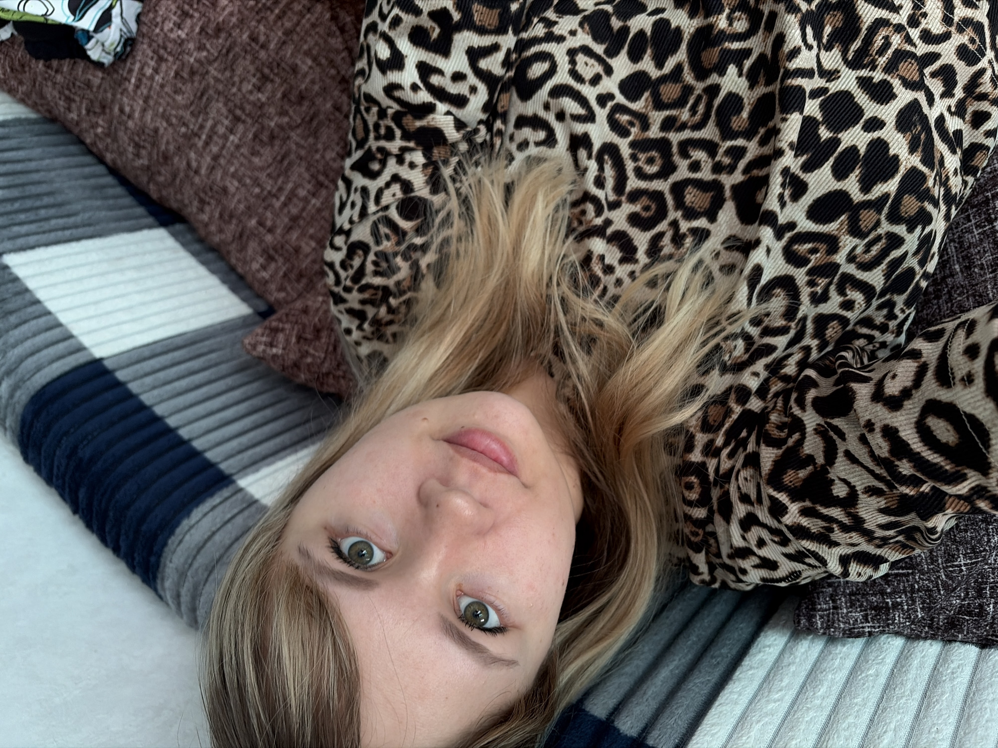A young woman with blonde hair and green eyes is lying on a bed or sofa. She is wearing a leopard-print top. The background includes a striped blanket, a brown cushion, and a small amount of fabric with a floral print. The location is Chaikovsky, Russia.
