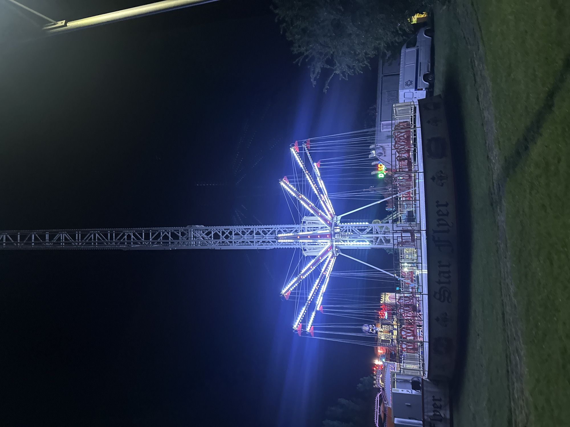This is a nighttime, eye-level, medium shot of a Star Flyer amusement ride in an outdoor setting, possibly a carnival or fair, at the city of Unknown, Unknown. The ride features a tall, white, metal tower structure with a spinning mechanism at the top. Attached to the spinning mechanism are multiple chains, each with a swing seat at the end. The entire ride is illuminated with bright white and blue lights. The base of the ride is circular and appears to be decorated with the words "Star Flyer." In the background, there are other amusement rides, buildings, and a white van. The sky is dark, indicating it is nighttime. The ground is covered in grass, and there is a tree on the right side of the frame.