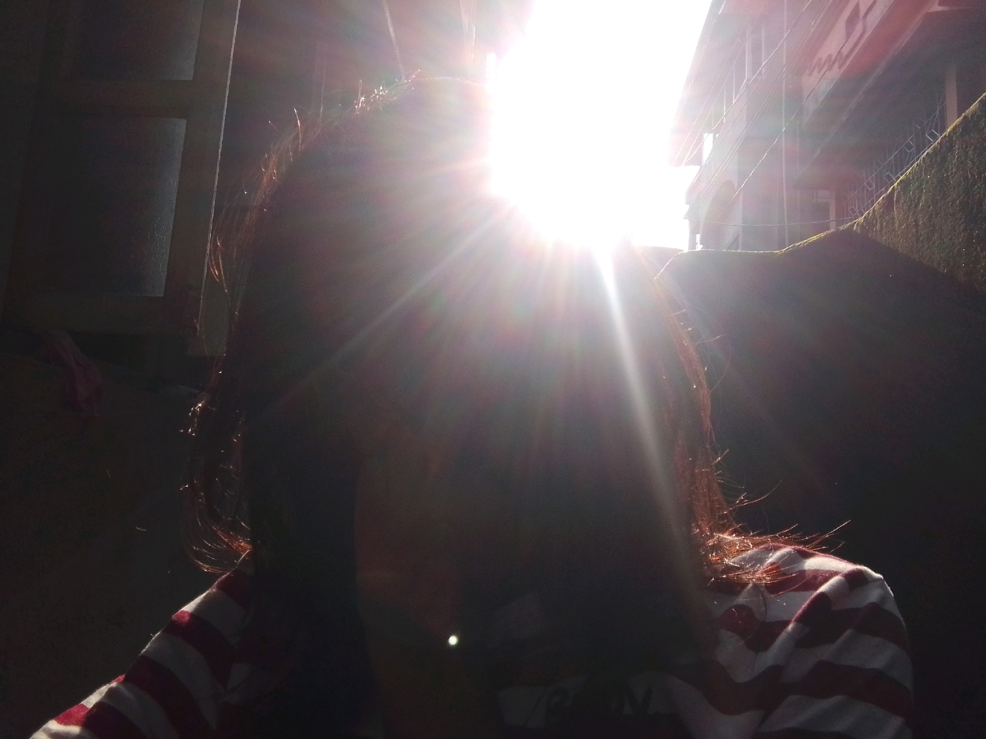 The image captures a person, partially obscured by bright sunlight, wearing a red and white striped shirt. The sun is positioned high in the frame, creating a lens flare that dominates the upper half of the image. In the background, a building with multiple stories and windows is visible, along with what appears to be a dark, textured wall or structure on the right. The lighting suggests it is daytime. The scene is consistent with an outdoor setting in Siliguri, India, possibly a residential area or a street.