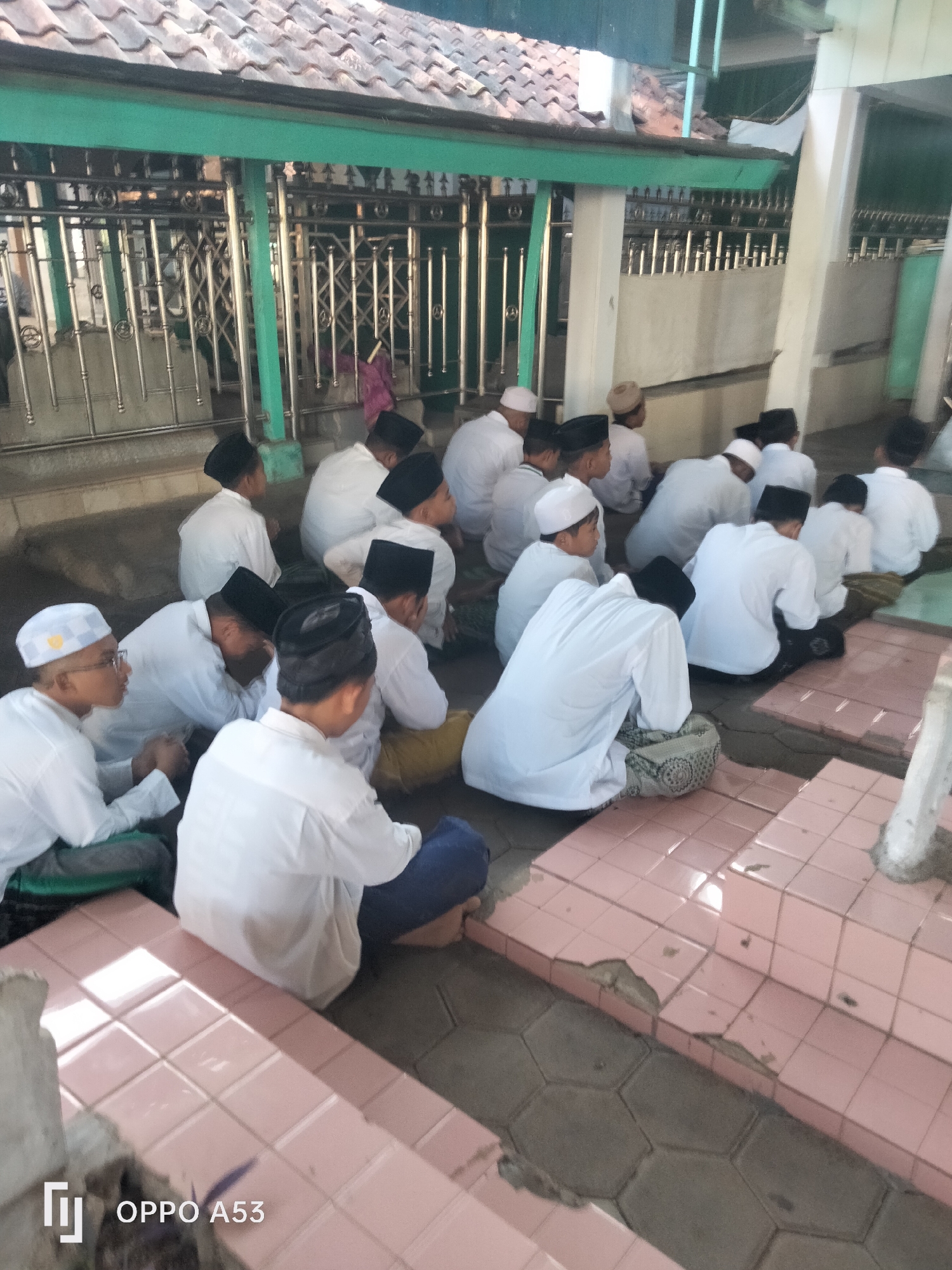 In Badung, Indonesia, a group of young boys are seated on the ground, facing away from the camera. They are dressed in white Islamic attire and black caps. Some are wearing glasses. They appear to be engaged in a religious activity. The setting seems to be an outdoor area with a tiled floor and a building with a tiled roof in the background. A metal fence is visible in the distance.
