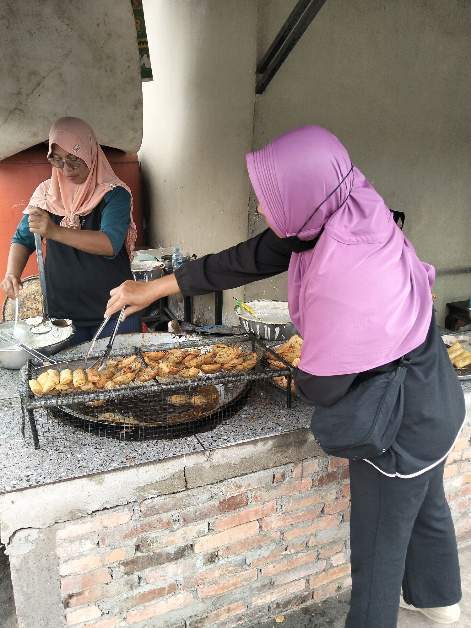 Two women are seen preparing food at an outdoor food stall in the city of Arang Limbung, Indonesia. The woman on the left, wearing a peach-colored hijab and glasses, is using a ladle to transfer fried items from a large metal bowl to a sieve. The woman on the right, wearing a purple hijab, is using tongs to flip pieces of food being fried in a large wok. The food appears to be battered and fried bananas. A metal rack holds more fried items above the wok. The stall is constructed with a brick base and a countertop. Behind the stall, a concrete wall and some metal structures are visible. The ground is covered with pebbles and a few fallen leaves.