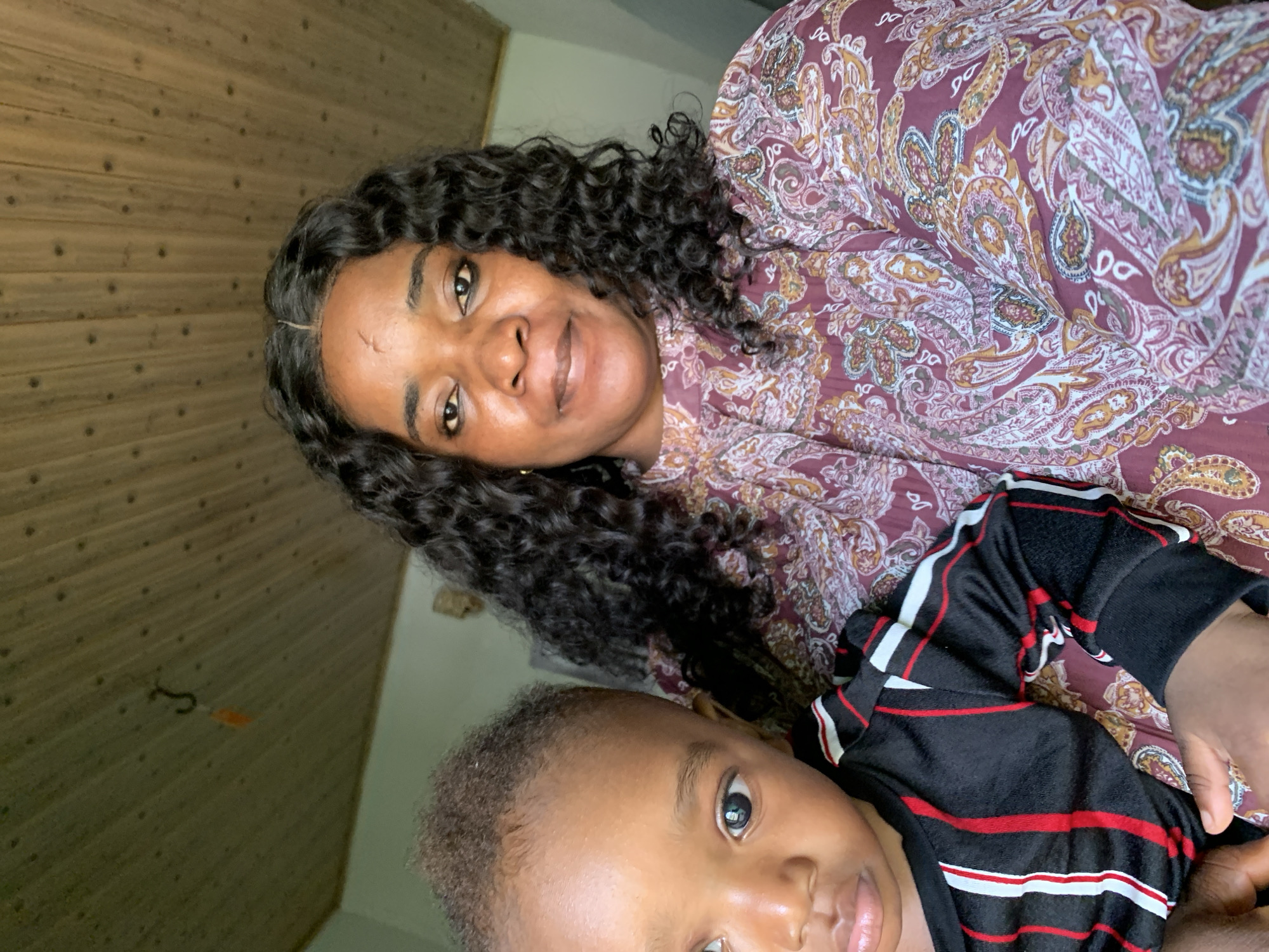 A woman and a young child are seated close together, with the woman on the right holding the child on her left. The woman has dark, curly hair and is wearing a long-sleeved, paisley-patterned top in shades of burgundy, pink, and yellow. She appears to be looking towards the camera. The child, facing left, has dark skin and short, curly hair. The child is wearing a black, long-sleeved shirt with red and white stripes. The background consists of a wood-paneled ceiling and a pale green wall. A small, dark hook with an orange object hanging from it is visible on the ceiling. The scene suggests an indoor setting, possibly a home. The specific location of Barakin Akawo, Nigeria, is not discernible from the visual information provided in the image.