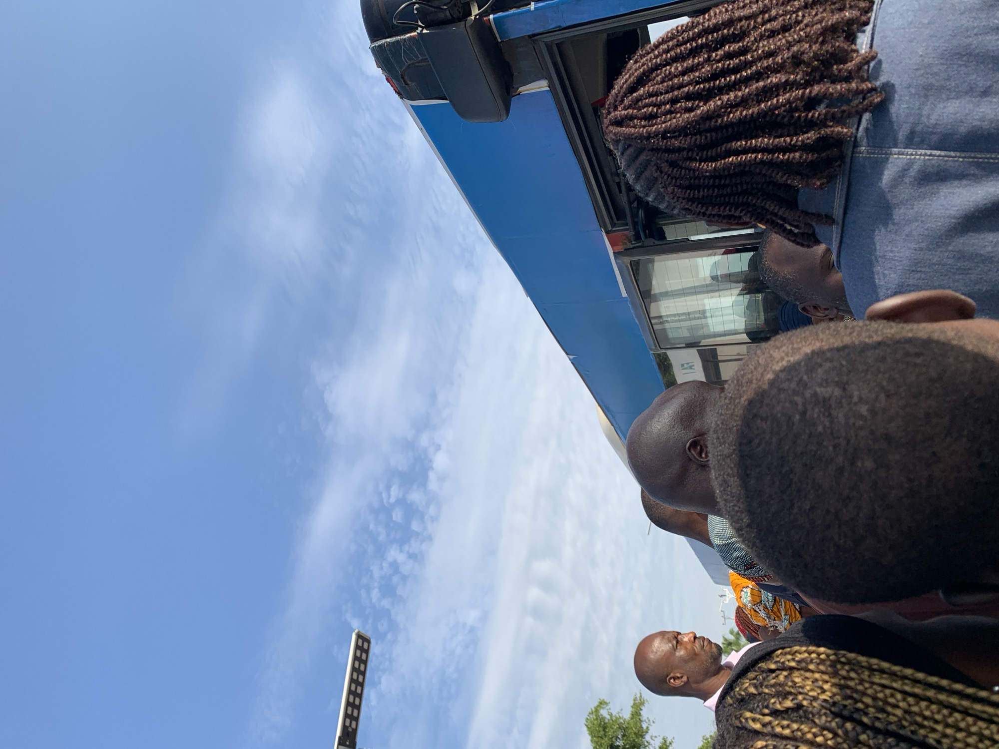 A group of people are standing in front of a blue bus, some of them appearing to be boarding. The scene is outdoors with a clear blue sky and some clouds. A street light is visible in the upper left corner of the image. The people are diverse in appearance, with varying hairstyles and clothing. The overall impression is that of a busy public transportation stop, possibly in a developing urban area. The location is identified as Rafin Par, Nigeria.