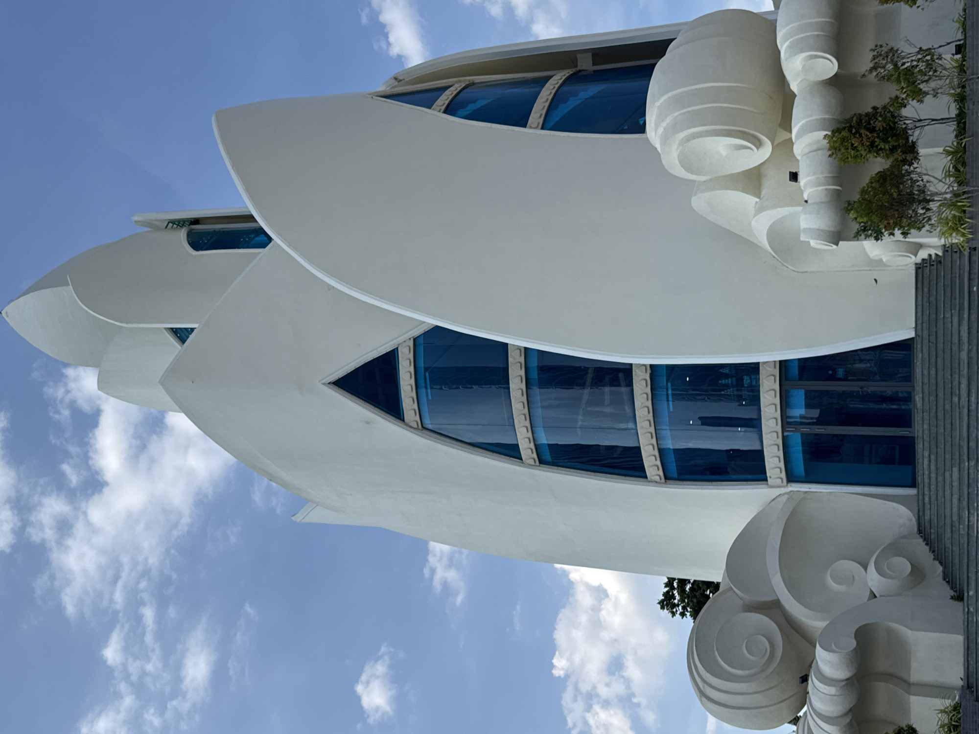 The image shows the exterior of the Long Son Pagoda, a Buddhist temple located in Nha Trang Ward, Vietnam. The pagoda features a distinctive white, multi-tiered structure with curved architectural elements resembling lotus petals. Large, arched windows with blue glass are visible on the facade. The base of the structure is adorned with white sculptural elements that appear to mimic swirling clouds or waves. In front of the pagoda, a set of gray stairs leads up to the entrance, which consists of double glass doors. Small trees and shrubs are planted along the base of the structure. The sky above is blue with scattered white clouds. No people are visible in the image.