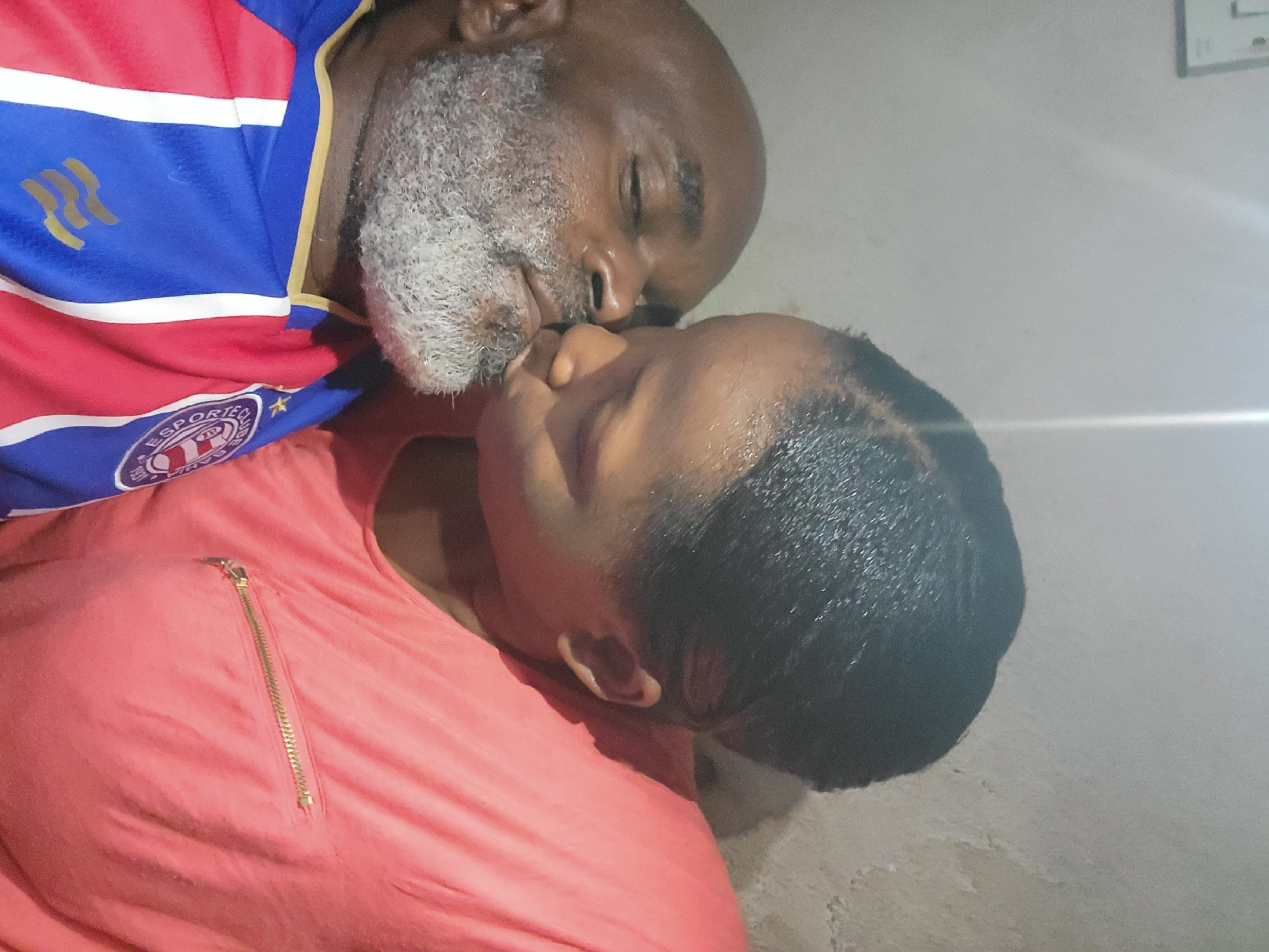 Two people are close together, possibly embracing or kissing. One person, wearing a blue and red sports jersey with a logo on the chest, has gray facial hair. The other person is wearing a salmon-colored shirt. The setting is indoors, with a white wall in the background. The location of the image is Sabon Tasha, Nigeria.