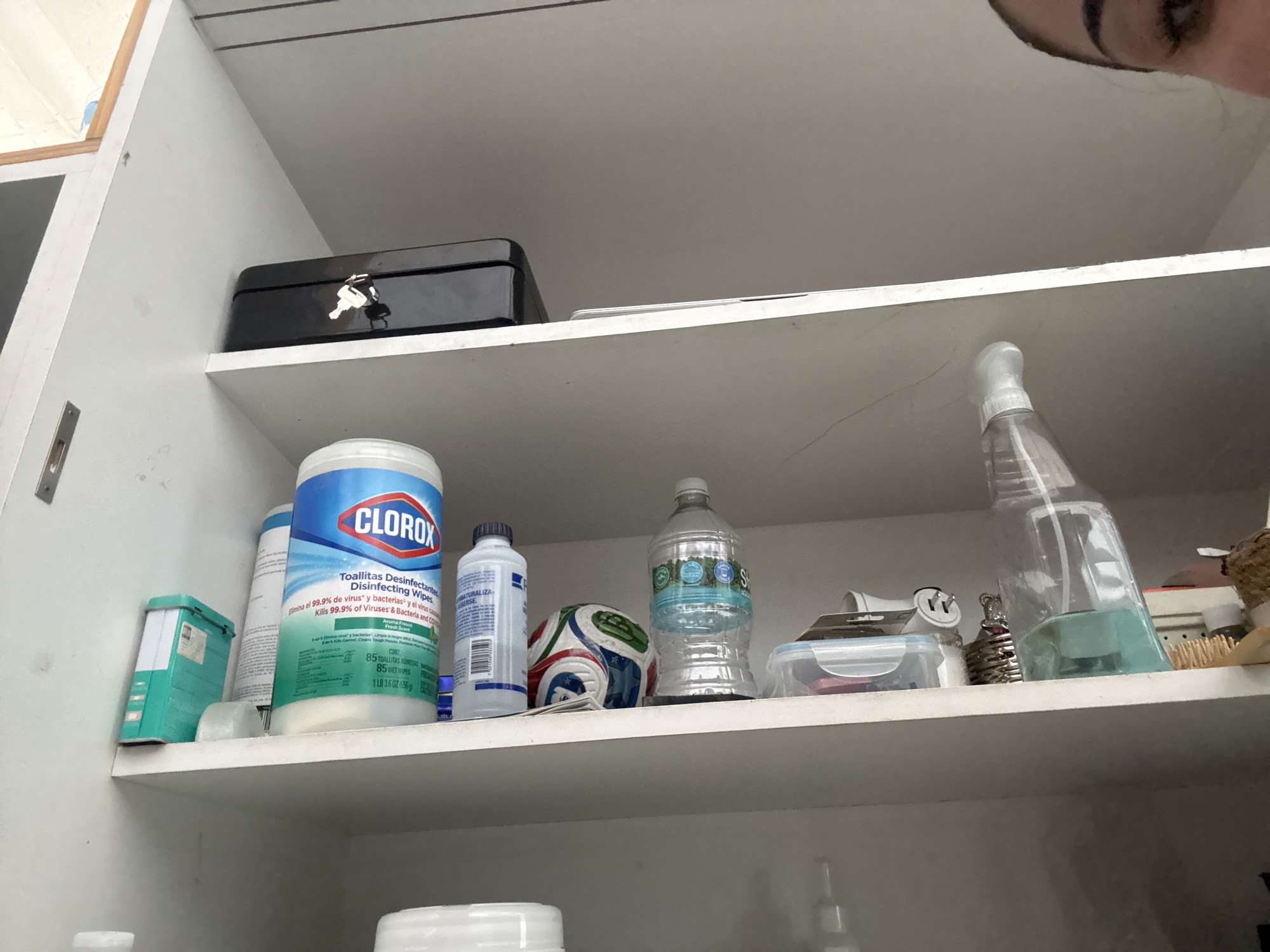The image captures the interior of a white cabinet with two visible shelves, viewed from a low, upward angle. In the top right corner, a portion of a person's eye and eyebrow is visible, suggesting someone is looking into or interacting with the cabinet.
On the upper shelf, a black metal box with a key in its lock rests on the left. To its right, a thin, light-colored object is partially visible.
The middle shelf holds an assortment of household items. Prominently featured on the left is a large container of Clorox Disinfecting Wipes. Next to it, a white bottle with a blue cap stands upright. A small, multi-colored (blue, white, and red) ball, resembling a mini soccer ball, sits between the bottles. Further right, a clear plastic water bottle is present, and behind it, a small clear plastic container with a grey lid. A clear spray bottle containing a light blue liquid is positioned on the far right of the shelf. Behind these items, a brush with wooden bristles, a silver coiled object, and a white electrical plug adapter are also discernible. On the far left of the shelf, a turquoise or teal rectangular organizer is partially visible behind the Clorox container.
Visible text on the Clorox container includes "CLOROX," "Toallitas Desinfectantes," "Disinfecting Wipes," "Elimina el 99.9% de virus y bacterias," "Kills 99.9% of Viruses & Bacteria," "Aroma Fresh Scent," "85 TOALLITAS HUMEDAS," "85 WET WIPES," and weight details. Partially visible text from other labels includes "ATURALIZA" and "EBERSE." The presence of Spanish text on the Clorox wipes, combined with the context of Puebla, Mexico, suggests a location where Spanish is spoken. The setting appears to be an indoor storage area, likely in a home.