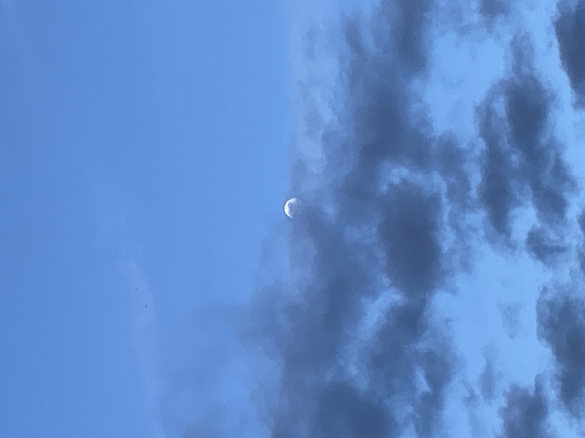 The image shows a daytime view of the sky, primarily featuring a light blue expanse with a crescent moon visible. Darker, textured clouds are present on the right side of the frame. The exact location in Budva, Montenegro cannot be determined without further information or context. There are no people, objects, actions, or interactions visible.