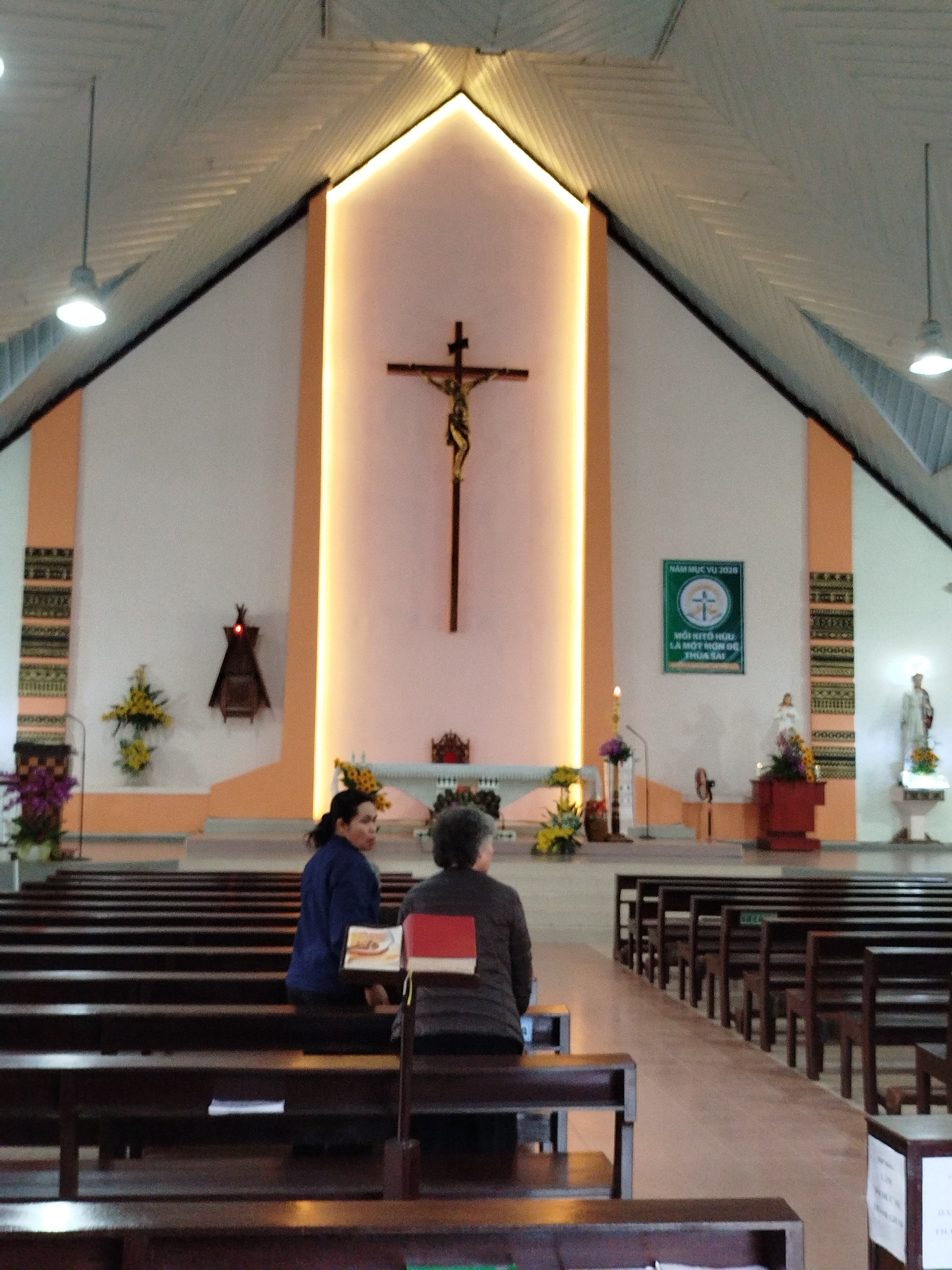 The image depicts the interior of a church located in K'Long, Vietnam. The church features a high, vaulted ceiling with exposed beams and pendant lighting. At the front of the church is a raised altar area with a crucifix as its central focus, flanked by floral arrangements, statues, and a banner in Vietnamese. Rows of wooden pews extend from the front to the back of the church. Two women are seated in one of the pews in the foreground, facing the altar. One wears a blue shirt, and the other a brown one, with books or pamphlets visible on the back of the pew in front of them. The overall setting suggests a place of worship during a quiet moment, possibly before or after a service.