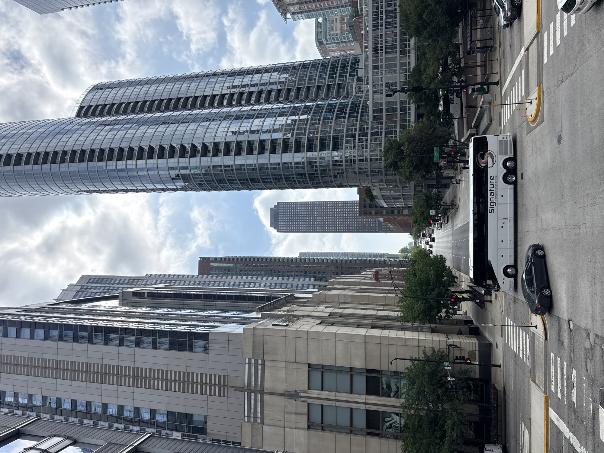 The image captures a street-level view of a city intersection in Chicago, Illinois, USA, characterized by towering skyscrapers and urban infrastructure. In the foreground, a white tour bus with "Signature Transportation Group" printed on its side is positioned in the middle of the intersection, having just passed a crosswalk. A dark sedan is in the process of turning, also having crossed a crosswalk. The intersection is marked with white lines and yellow bollards. Traffic signals are visible, with one showing a red light.
The environment is dominated by modern high-rise buildings. To the left, a light-colored building with large windows and vertical detailing rises high, featuring a sign with what appears to be artwork. Further down the street, another tall building with a textured facade is visible. On the right side of the image, two prominent, curved skyscrapers with numerous balconies and reflective glass facades dominate the skyline. These buildings are interspersed with smaller buildings and trees lining the sidewalks.
In the background, more tall buildings stretch towards the sky, and a multi-lane street with several vehicles can be seen moving away from the viewer. The sky is partly cloudy, with patches of blue sky visible through the white clouds. The overall impression is a dynamic urban scene in a bustling metropolitan area.