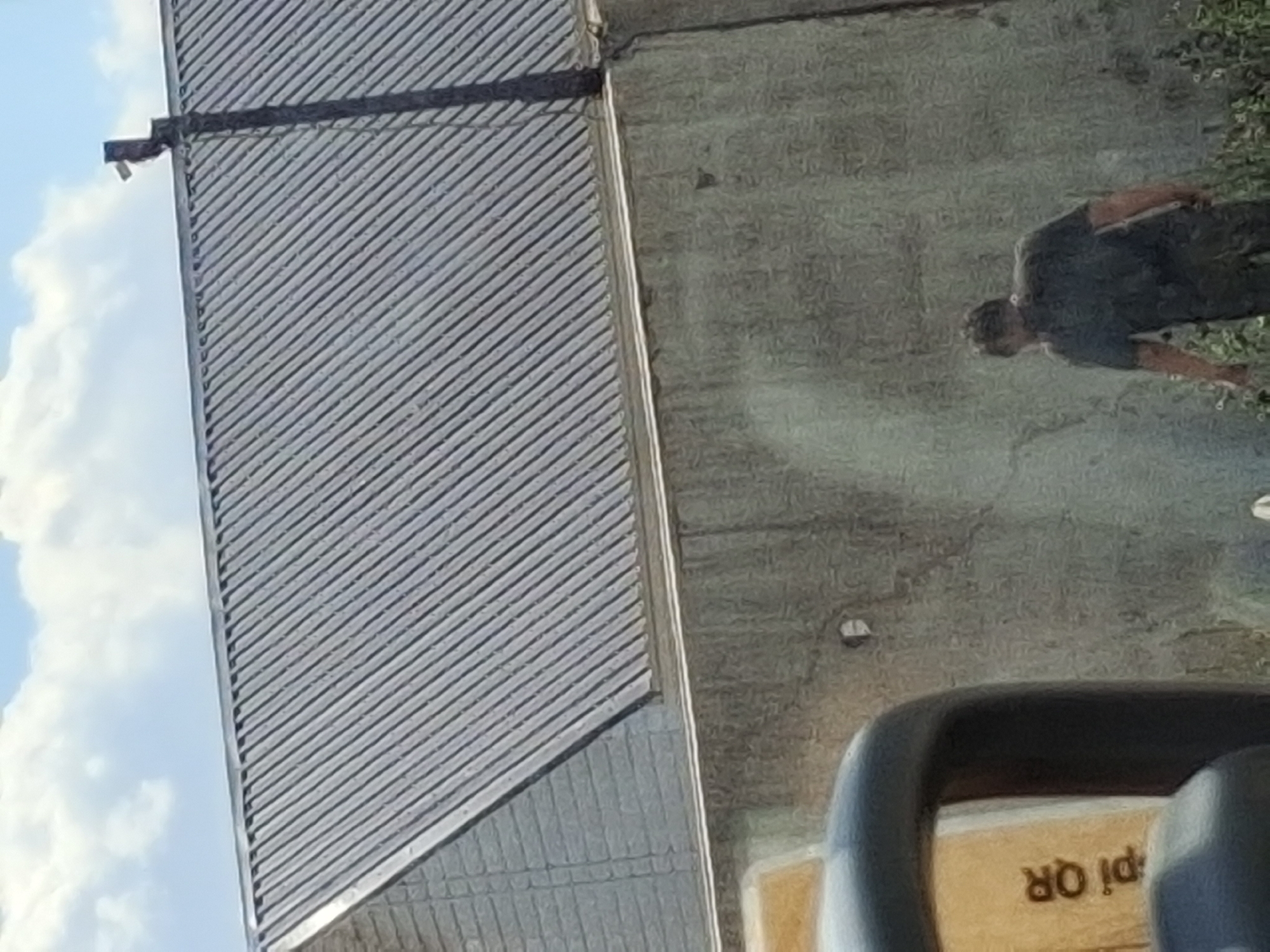 The image shows a person in a dark t-shirt and pants standing near a concrete wall. The person appears to be outdoors, with a corrugated metal roof visible above the wall and a cloudy sky in the background. A part of a vehicle, possibly a car, is visible in the foreground on the left side of the image, with a partial view of a yellow sign that includes the text "pi QR". There are some plants visible at the bottom right of the image. The overall scene appears to be in an industrial or urban setting. The provided context mentions the city of Semey, Kazakhstan, but there are no specific visual cues in the image to confirm this location.
