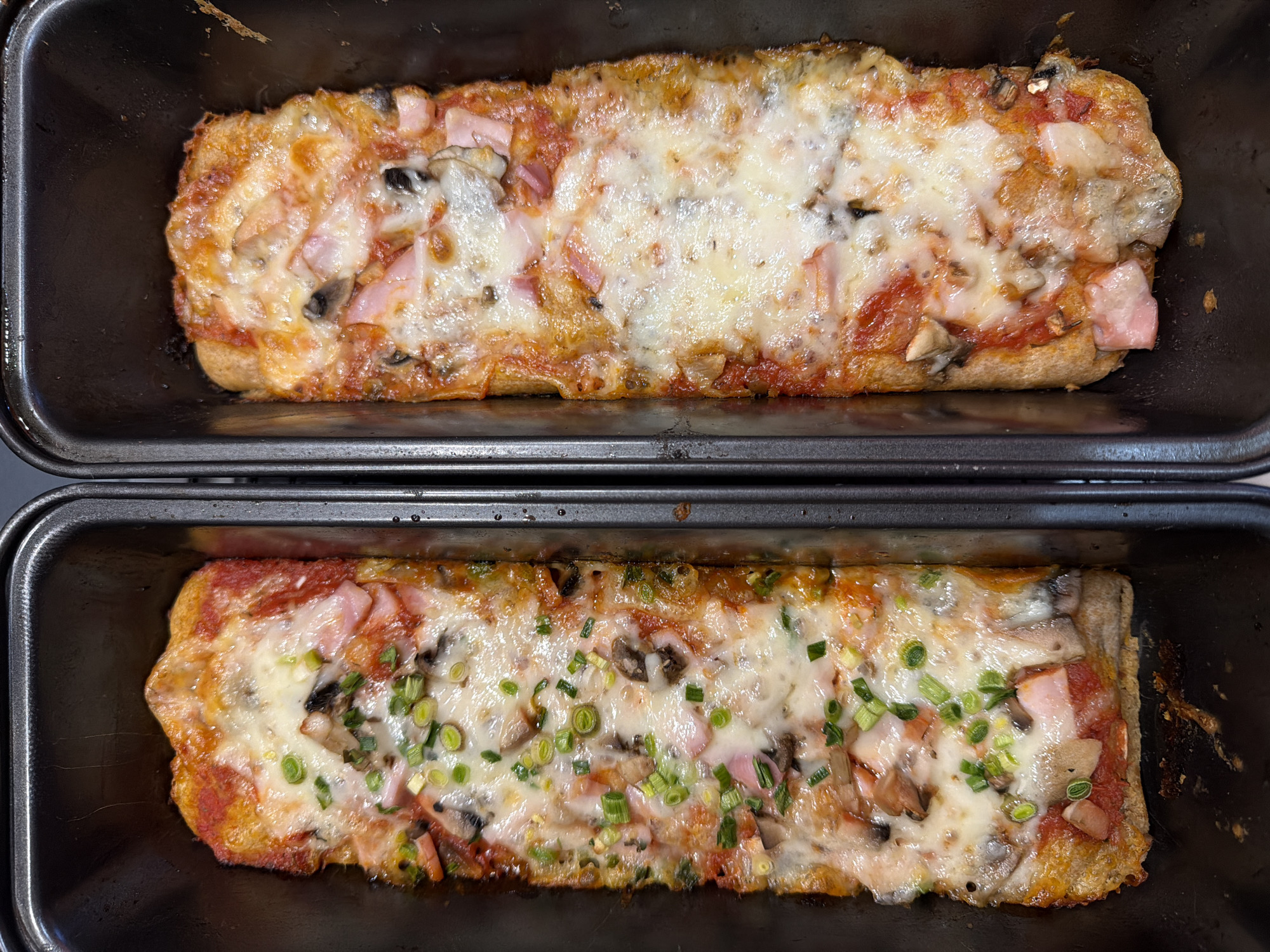 The image shows two rectangular pizzas, each in its own dark metal baking pan. The pizzas appear to have a similar base with toppings of tomato sauce, cheese, mushrooms, and ham. The lower pizza has the addition of chopped green onions or chives scattered across the cheese. The baking pans are stacked, with the pan containing the top pizza resting on top of the other. The image does not provide information about location or the city of Varna, Bulgaria, nor does it depict any people or specific actions/interactions.