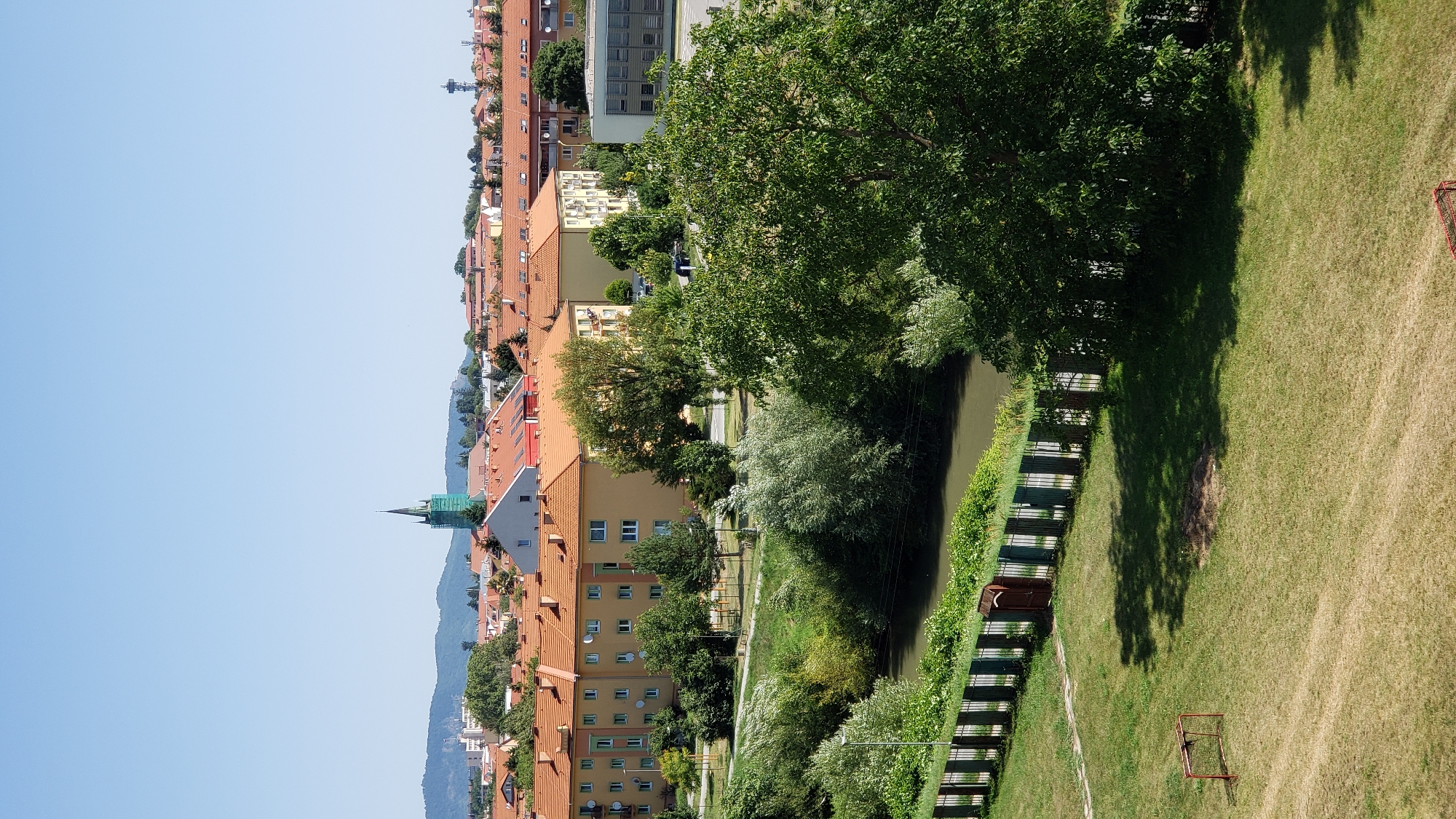 This is a daytime, eye-level, vertical, exterior, full shot of a residential area in Prešov, Slovakia. The scene is dominated by several apartment buildings with orange-tiled roofs and yellow facades, interspersed with trees and greenery. A river runs through the middle ground, bordered by a fence and more vegetation. In the background, a church steeple, visible from the crops, is partially covered in green scaffolding. The sky is clear and blue, with a few distant hills or mountains on the horizon. In the foreground, a grassy area contains a small red soccer goal and some patchy dry grass.