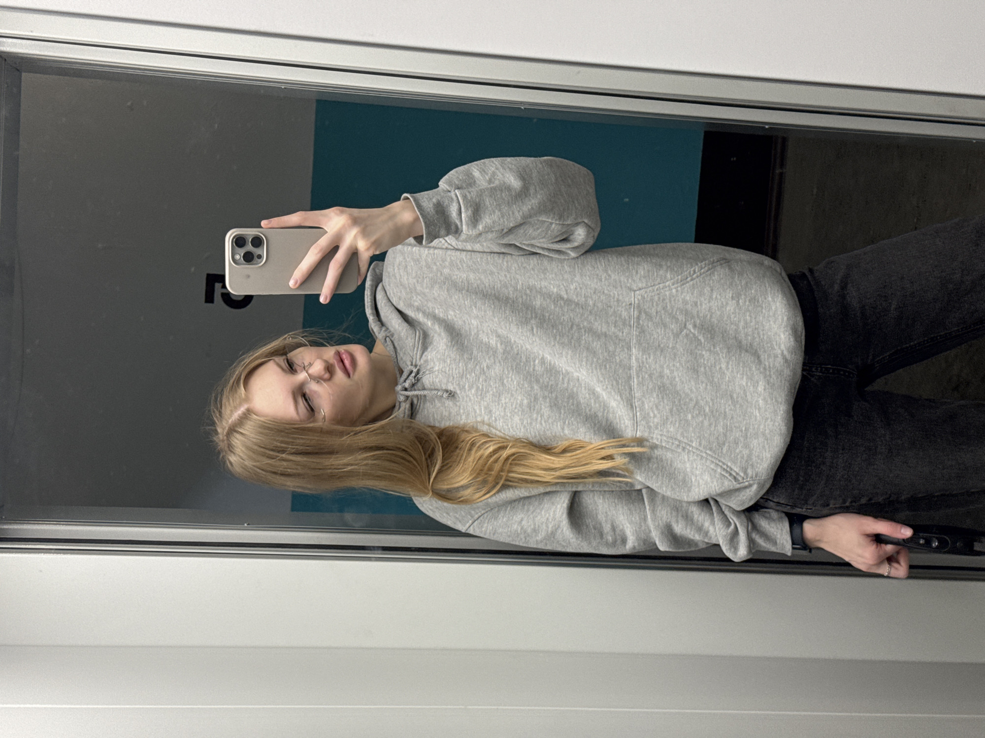 A young woman with blonde hair, wearing a grey hoodie and dark jeans, takes a selfie in a mirror. She is holding a smartphone in her right hand, angled to capture her reflection. Her hair is long and wavy, falling over her shoulder. She appears to be wearing glasses and a nose ring. The background shows a wall painted in a teal color and a dark doorway. There is some faint, dark text on the teal wall that is not clearly legible. The lighting suggests it could be daytime, possibly indoors in a well-lit area, or outdoors in diffused light. The overall mood is casual and everyday.