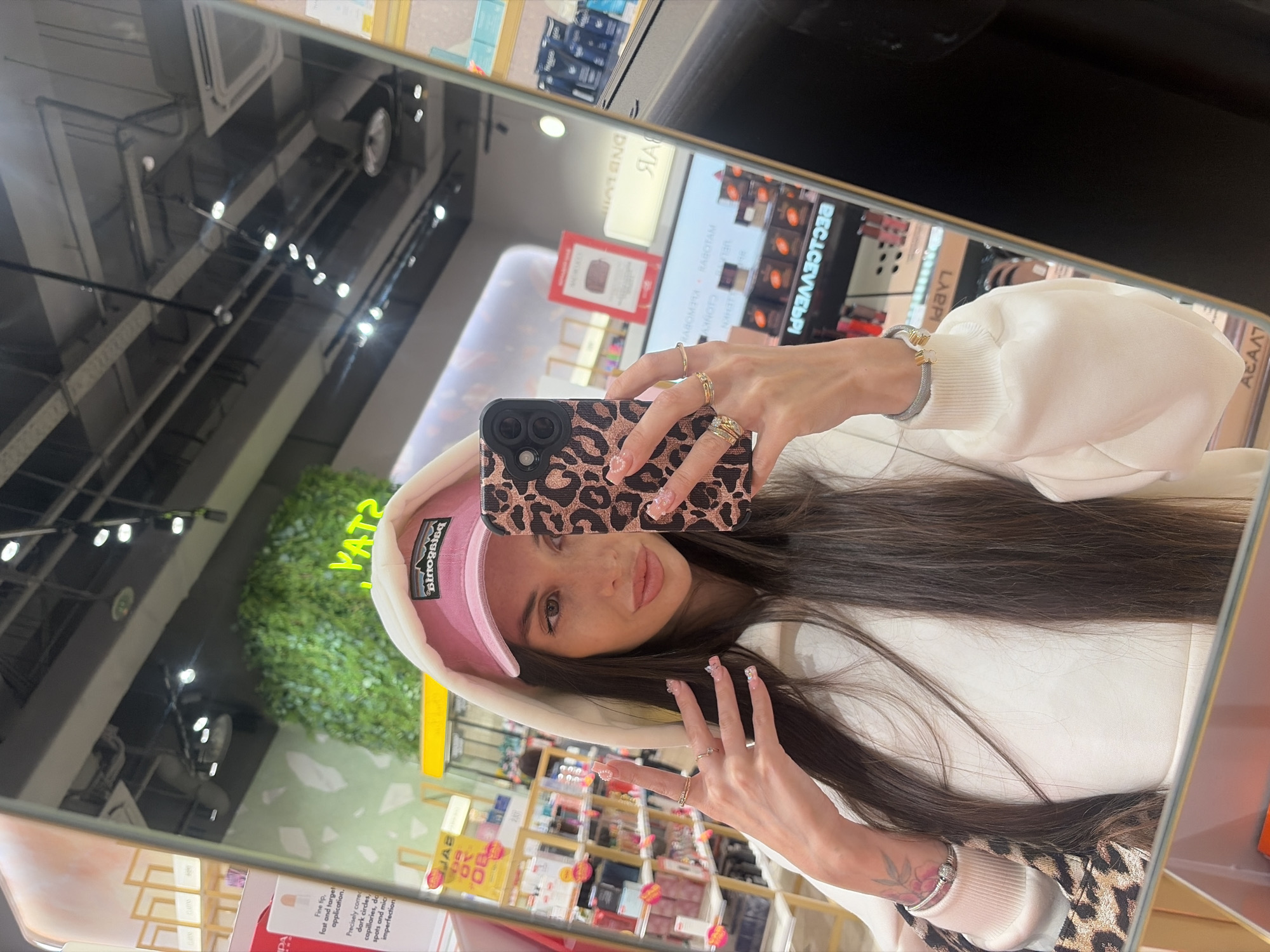 A young woman takes a mirror selfie in what appears to be a retail store, likely a cosmetics or beauty shop, located in Izhevsk, Russia. She has long, dark brown hair and is wearing a white hooded sweatshirt. A pink baseball cap with the "Patagonia" logo is visible under her hood. Her phone, which has a leopard print case, is held up to capture her reflection. She is looking directly at the phone's camera with a slight smile.
Notable details include several rings on her fingers and a silver-colored bracelet with a floral charm on her left wrist, where a faint tattoo is also visible. Her nails are manicured with light-colored polish and small decorative elements.
The background, visible in the mirror's reflection, shows an indoor environment with overhead track lighting and industrial-style exposed pipes on the ceiling. Shelves stocked with various products are evident, along with several display stands. A green wall feature, possibly artificial foliage, is seen on the left.
Visible text and signs, some reversed due to the mirror reflection and some in Cyrillic script, include: "Patagonia" (on her cap), "YATZ", "EHEN", "CJONKY", "RABO OTAM", "DND L", "ЯА", "RECICEVVELPI", "LAPPI", "ΑΕΑΛΙ", "Білозвіва", and "KLE WORVE". A product description on a sign mentions "Fine lip, fast and target application. Precisely correct dark circles, capillaries, dark spots and minor imperfection," and the word "GLOW" is seen on another stand. The presence of Cyrillic text aligns with the given location context of Izhevsk, Russia.