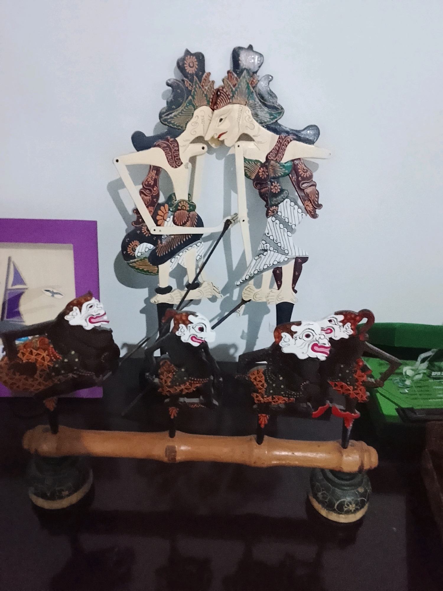 The image displays a collection of Wayang Golek, traditional Indonesian rod puppets, displayed on a dark, reflective surface, possibly a table. Two larger, elaborately dressed puppets stand upright in the background, facing each other, their hands extended. In the foreground, three smaller puppets are arranged on a light brown wooden beam supported by decorative dark bases. These smaller puppets are depicted with exaggerated facial features, including wide eyes and open mouths with red lips. They are dressed in dark fabrics adorned with orange and brown patterned sashes. To the left of the puppets, a framed picture with a purple border is partially visible, showing a sailboat and birds. The background is a plain, light-colored wall. The scene suggests a display of cultural artifacts, likely in a home or a cultural exhibition space in Slawi, Indonesia.