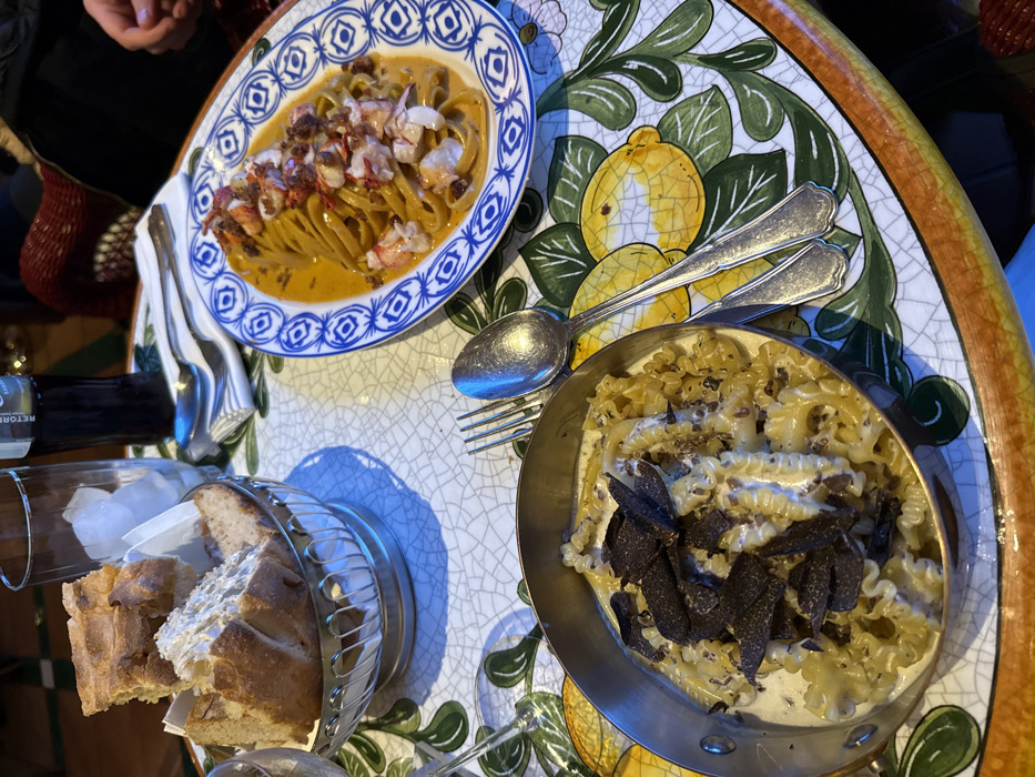 This image depicts a detailed view of a dining table in an indoor restaurant setting in Barcelona, Spain.
The table features a distinctive ceramic tile-like surface patterned with yellow lemons and green leaves on a white background with a crackle finish. Two main pasta dishes are presented:
1. On the left, a small, round, silver-colored pot contains a dish of mafalde or reginette pasta in a creamy white sauce, generously topped with thinly sliced black truffles. A silver fork and spoon are positioned to its right.
2. On the right, an oval plate with a blue and white geometric border holds tagliatelle or fettuccine pasta in an orange-hued sauce, garnished with visible chunks of white meat, consistent with lobster, and a dark, crumbled topping.
Other items on the table include a silver basket holding several torn pieces of crusty bread, lined with white paper, located in the upper left. Next to the bread, a tall, clear glass filled with ice cubes and a dark brown bottle with the "RETORNA" label visible are present. A neatly folded white napkin containing a fork and spoon rests between the bread basket and the oval pasta plate. A wine glass is partially visible on the far left edge of the frame.
A human hand, with light skin, is partially visible in the upper right corner of the frame, indicating the presence of at least one diner. The background is softly blurred and dimly lit, revealing a red woven texture, likely part of a chair or booth. The scene captures a meal ready for consumption.