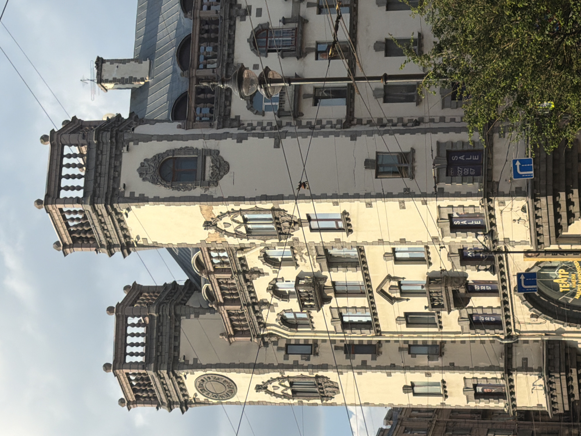 This is a photograph of a building facade that appears to be of historical or architectural significance. The building is tall and ornate, with multiple stories, decorative stonework, and numerous windows. The style of the architecture suggests it might be from the late 19th or early 20th century. Two prominent towers with intricate detailing and what look like balustrades flank a central section of the building.
Visible elements include:
* **Architecture:** The building has a light-colored facade with dark gray or black decorative trim. The towers have crenellated tops. There are arched windows and smaller rectangular windows, some with decorative surrounds.
* **Objects:** Several signs are visible. One sign appears to say "SALE" in large letters. Another sign, with text in Cyrillic script, is partially visible on a curved section of the building, and the word "ТЕАТР" (Theater) is discernible. Electrical wires and a lamppost with two globes are also present, crisscrossing the facade. A blue directional sign with a white arrow is on the right side of the image. A tree with green leaves is to the right of the building.
* **Scene:** The photograph is taken from a low angle, looking up at the building. The sky is visible in the background, suggesting it's daytime. The presence of overhead wires and the lamppost indicates an urban setting. The lighting suggests it is likely daytime, possibly late afternoon given the angle of the light.
* **Text:** "SALE" is clearly visible on at least two signs. Cyrillic text, including "ТЕАТР", is also present.
The overall impression is one of an old, established building in a city, possibly a commercial district or an area with cultural institutions like a theater. There are no people visible in the frame. The image appears to be taken on a clear day.