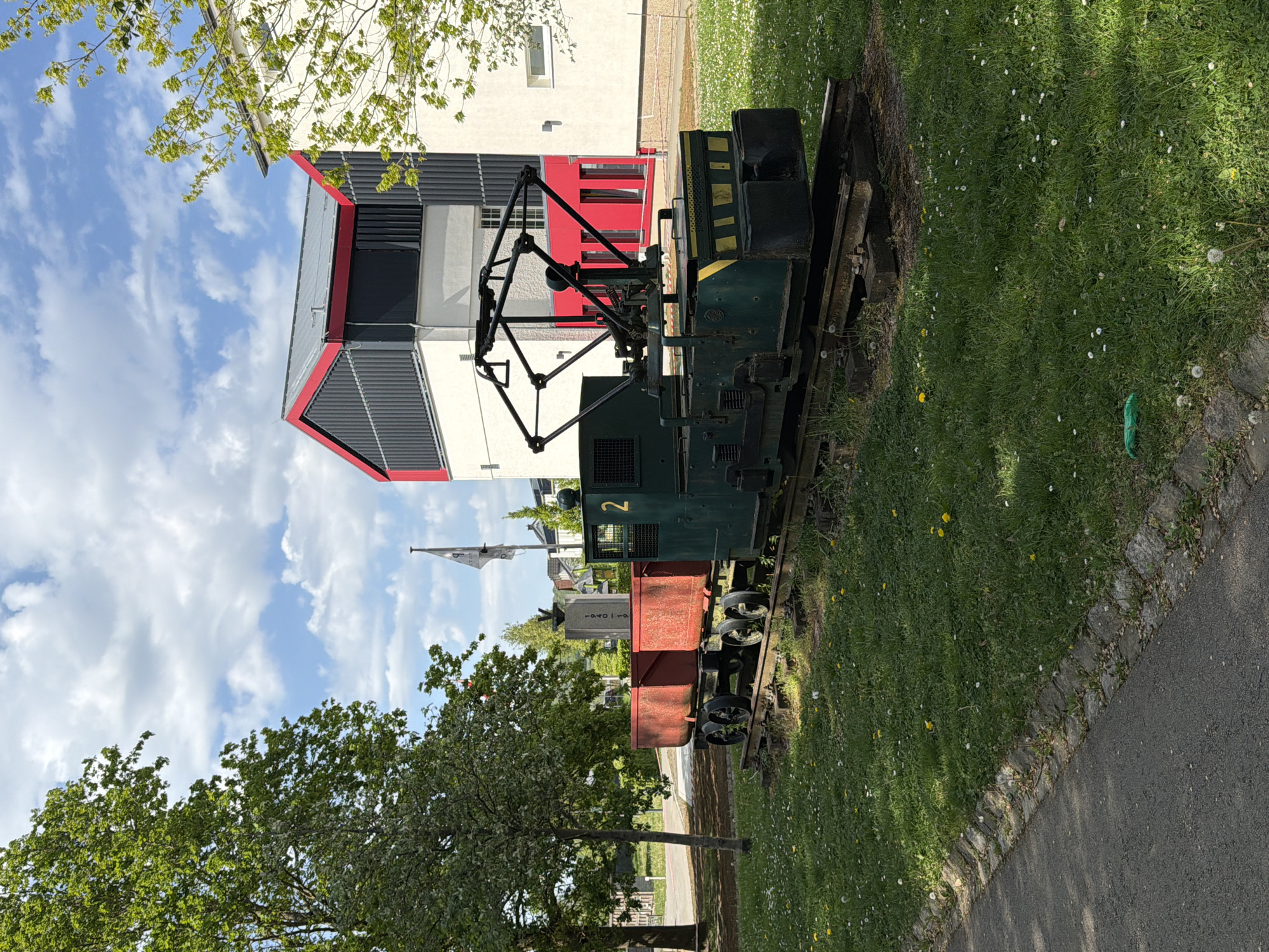 This image, taken in Pétange, Luxembourg, depicts a static display of an industrial locomotive and a small wagon set on tracks, situated in a grassy area next to a modern building.
The central subject is a dark green locomotive, number "2" painted in yellow on its side, featuring an intricate black pantograph-like structure on its roof. Attached to it is a rust-red, open-top wagon. Behind the wagon, a dark grey or black rectangular sign or marker is visible, with faint white text that appears to include "1940-19". The locomotive and wagon are positioned on a short section of railway track, which is embedded in well-maintained green grass. The grass is dotted with numerous small white and yellow flowers, resembling daisies and dandelions.
Adjacent to the grassy display area, a paved pathway runs along the right side of the frame. In the background, a striking modern building with a multi-layered design dominates. It features white walls, dark grey or black slatted panels, and prominent red structural or decorative elements, particularly on its roofline and around some window areas. Trees with vibrant green leaves frame the scene from the foreground and background, suggesting a spring or early summer setting.
The sky is bright blue with scattered fluffy white clouds, indicating clear and sunny weather. The overall impression is of a historical railway exhibit placed in a contemporary public space or campus-like environment. No people are visible, and there is no active event taking place, suggesting the scene captures a tranquil moment at a permanent outdoor display.